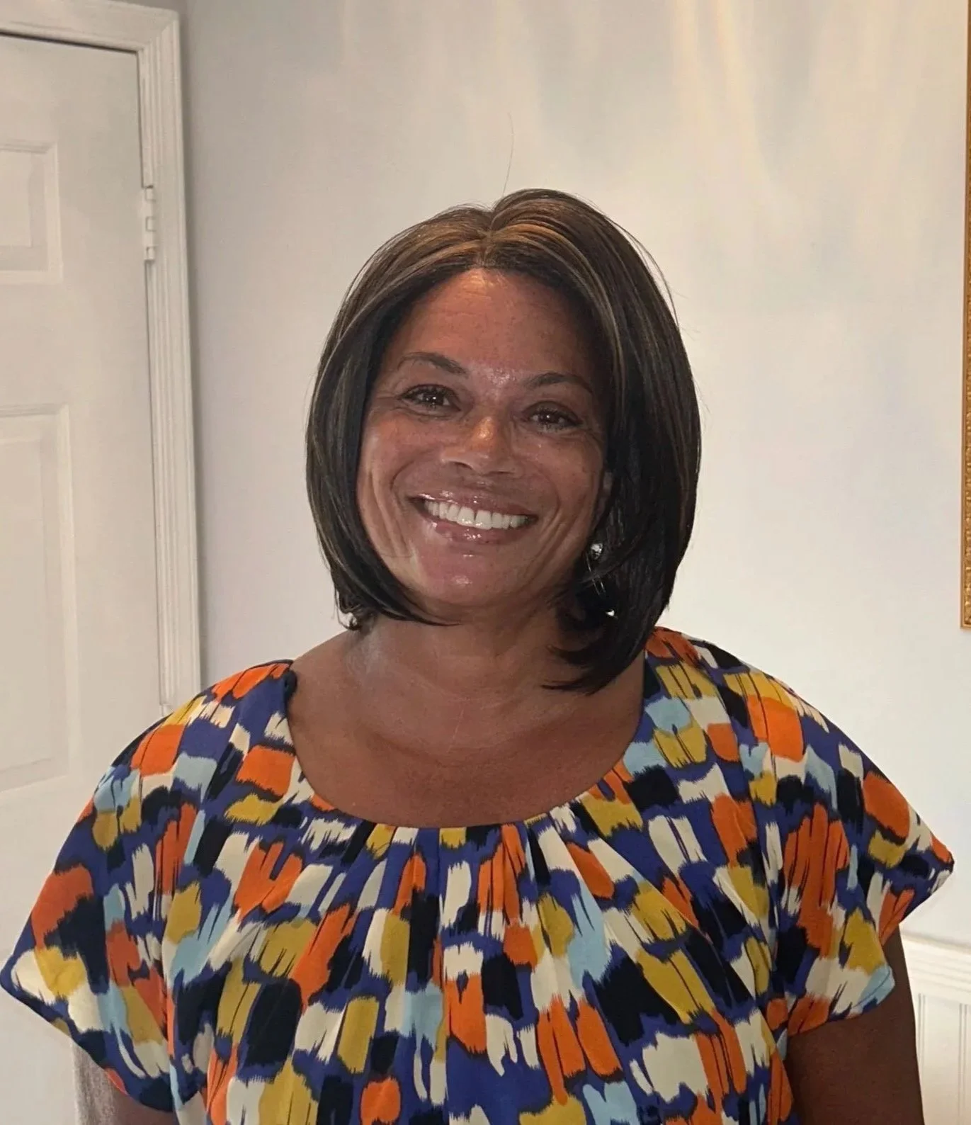 A woman with medium-length dark brown hair with highlights, smiling, wearing a colorful patterned blouse with orange, blue, yellow, and black. She is indoors against a white wall with a partially visible door on the left.