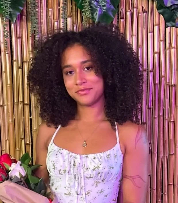 A woman with curly hair wearing a white floral dress, standing in front of a bamboo backdrop with colorful lighting, holding a bouquet of flowers.