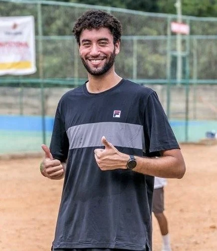 Smiling man with curly hair and a beard giving two thumbs up on a tennis court.