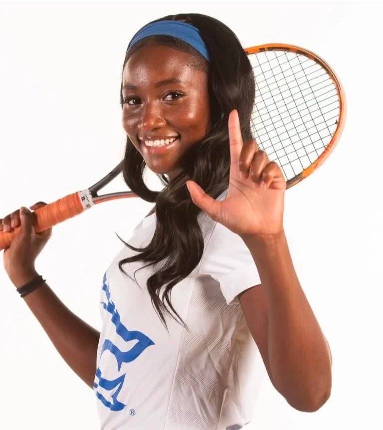 A young woman smiling, holding a tennis racket over her shoulder, wearing a white sports shirt with a blue logo, and a blue headband.