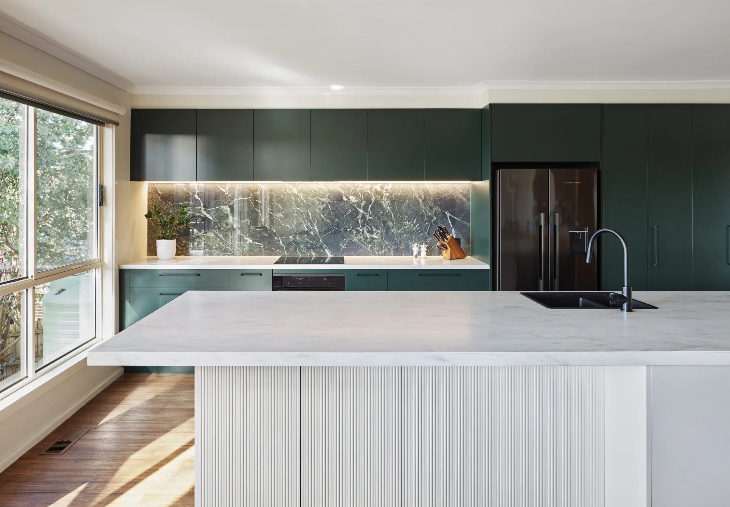Modern kitchen with green cabinetry, marble backsplash, white island with a black sink, faucet, and a knife block, next to large window letting in natural light.