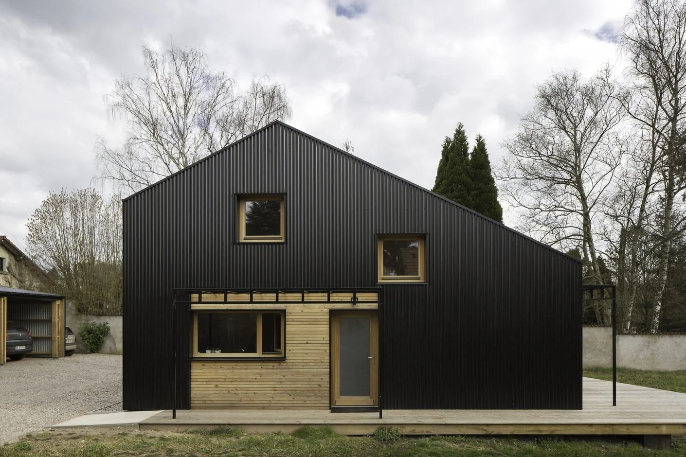 Black modern house with wooden accents and a sloped roof, surrounded by leafless trees and a gravel driveway.
