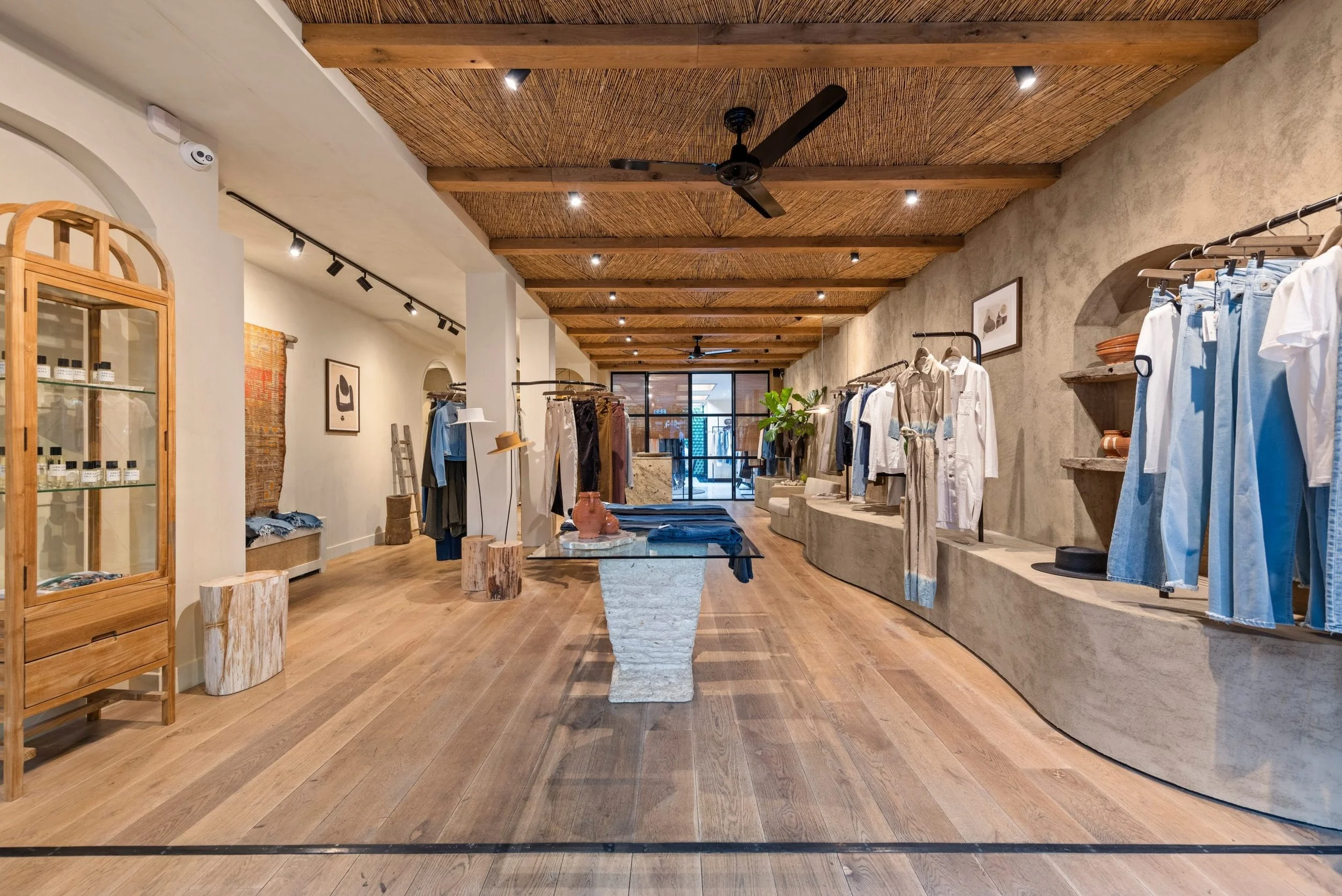 Interior of a retail clothing store with racks of clothes, wooden and stone display tables, plants, art pieces, and a wooden ceiling with a fan.