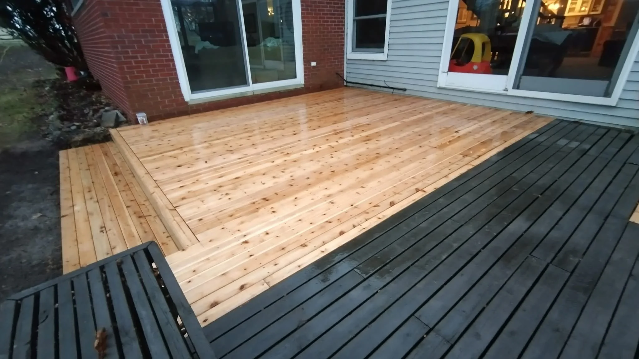 Newly installed wooden deck with a light finish outside a house, with a darker wood section adjacent, and large sliding glass doors leading inside.