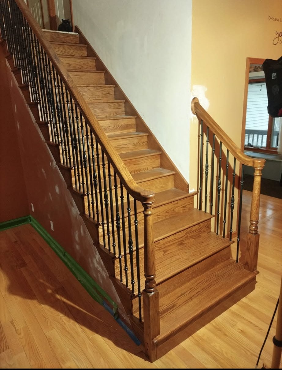 Interior of a house featuring a wooden staircase with black metal balusters and a rounded wooden handrail. The stairs are made of polished wood. To the right, there is a partial view of a room with a window and a door. The floor is made of wooden planks.
