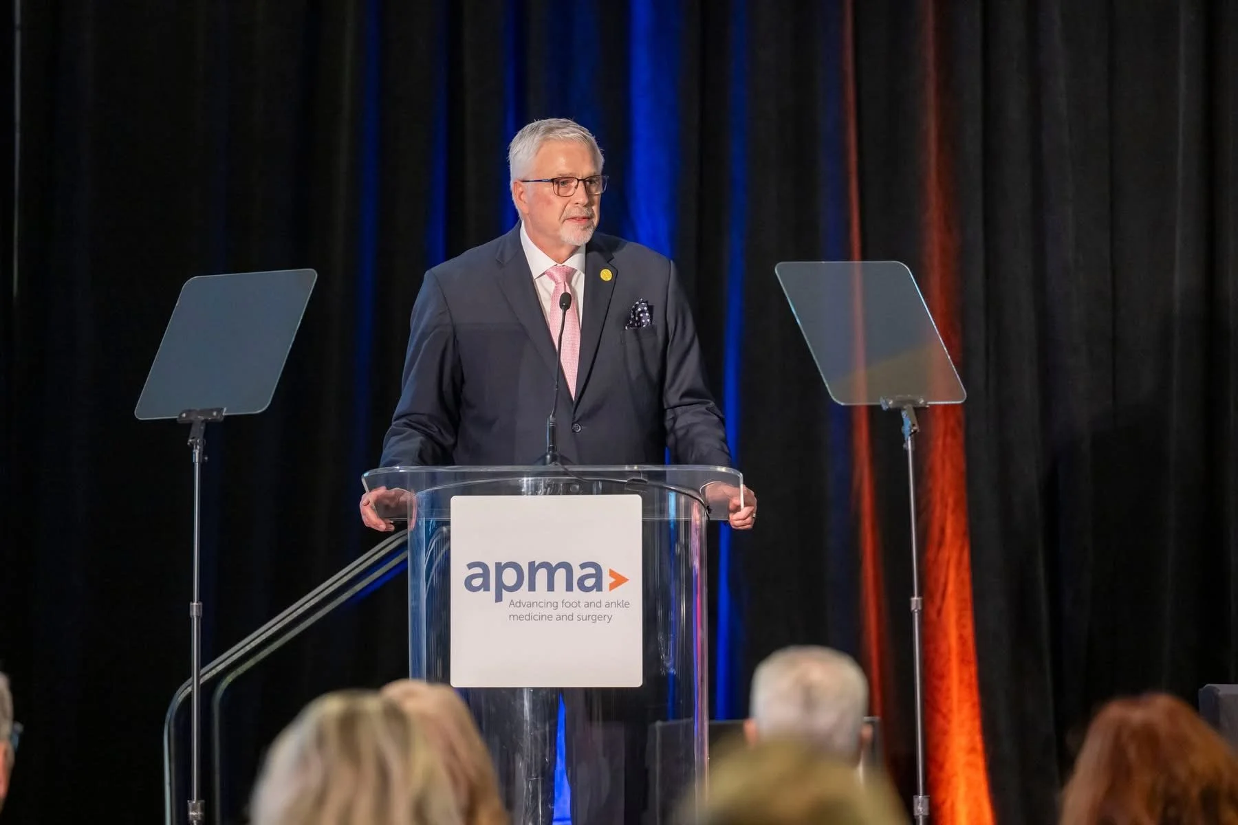 A man with gray hair and glasses speaking at a podium during a conference, with a black curtain background, two transparent teleprompters on either side, and the APMA logo on the front of the podium.