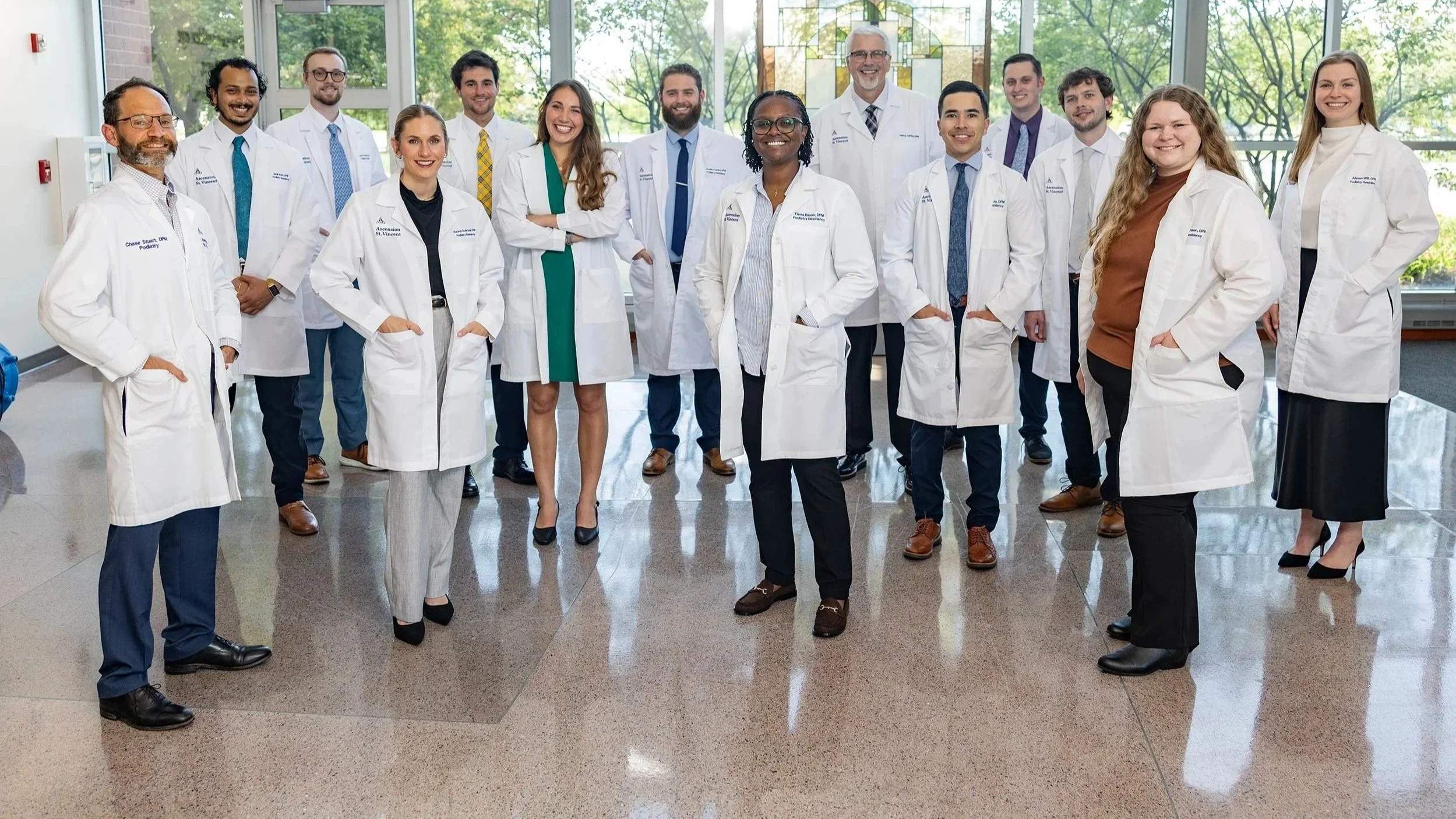 Group of diverse medical professionals and doctors standing together in a modern hospital lobby with large windows and natural light, smiling at the camera.