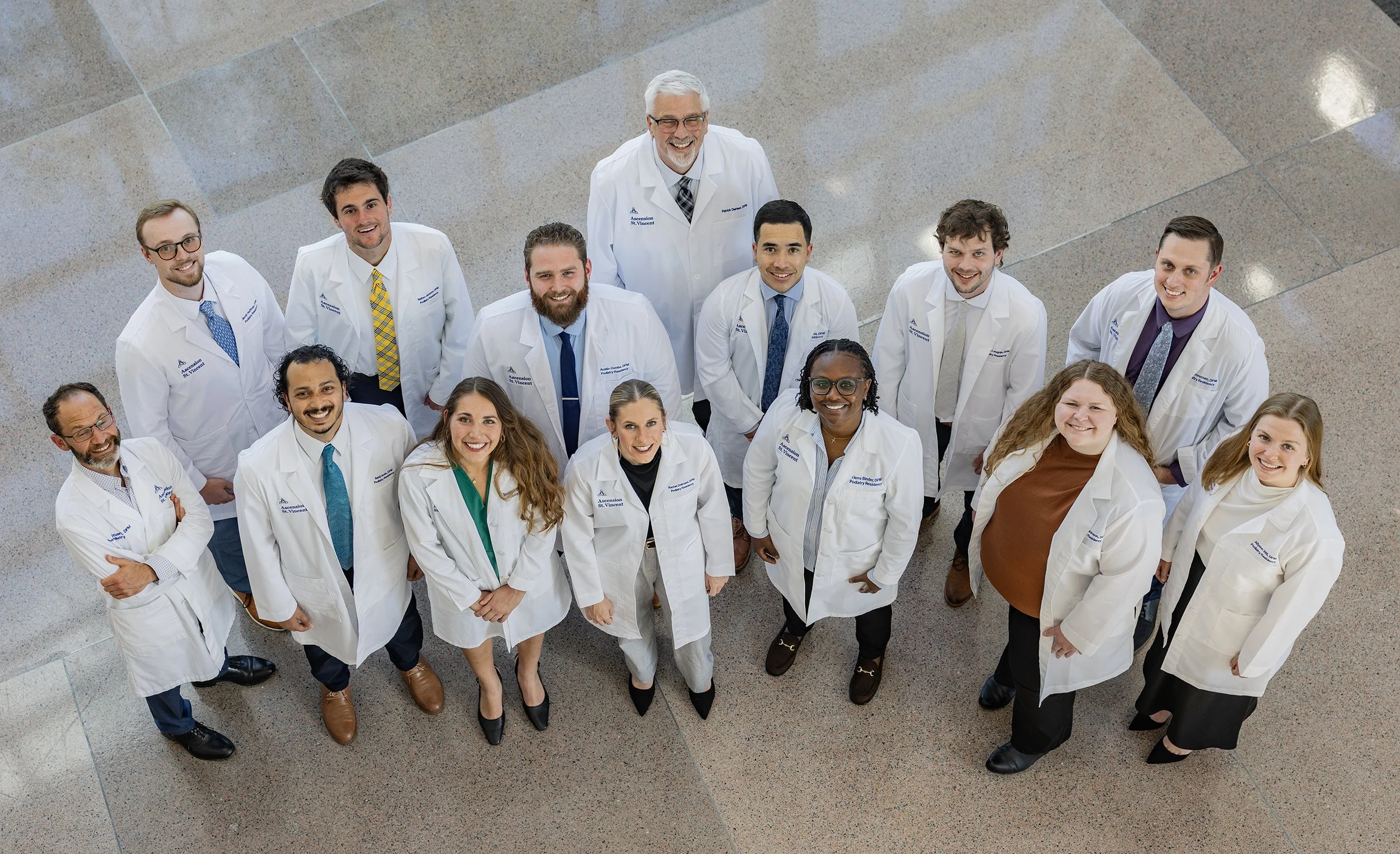 A group of people, likely medical professionals or students, is standing and looking up at the camera in a brightly lit indoor setting. They are wearing white lab coats and are smiling.