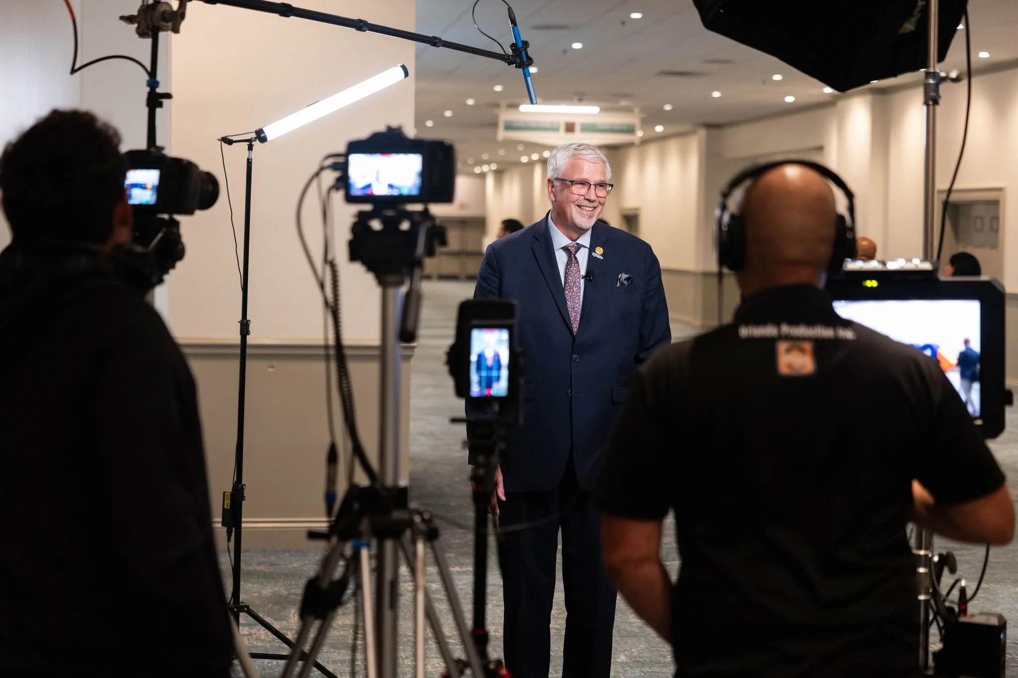 An older man with gray hair and glasses wearing a dark suit and tie is smiling and speaking during an interview or recording session in a conference hall, with professional camera crew and equipment capturing his image.