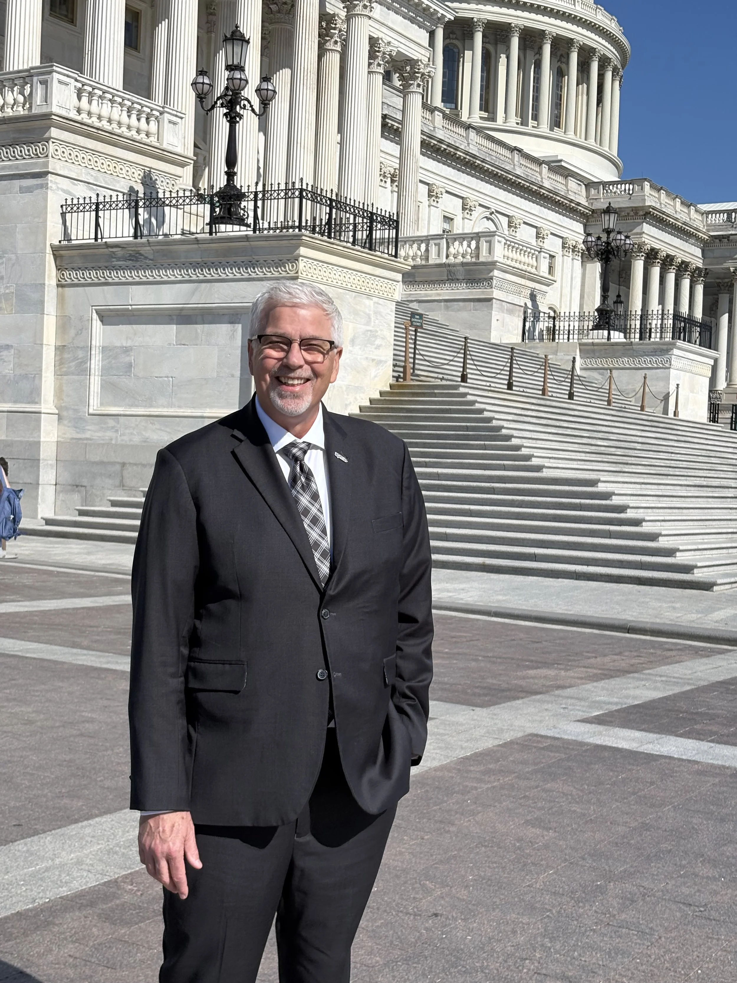 A man in a suit and glasses standing in front of the United States Capitol building with stairs and decorative lampposts.