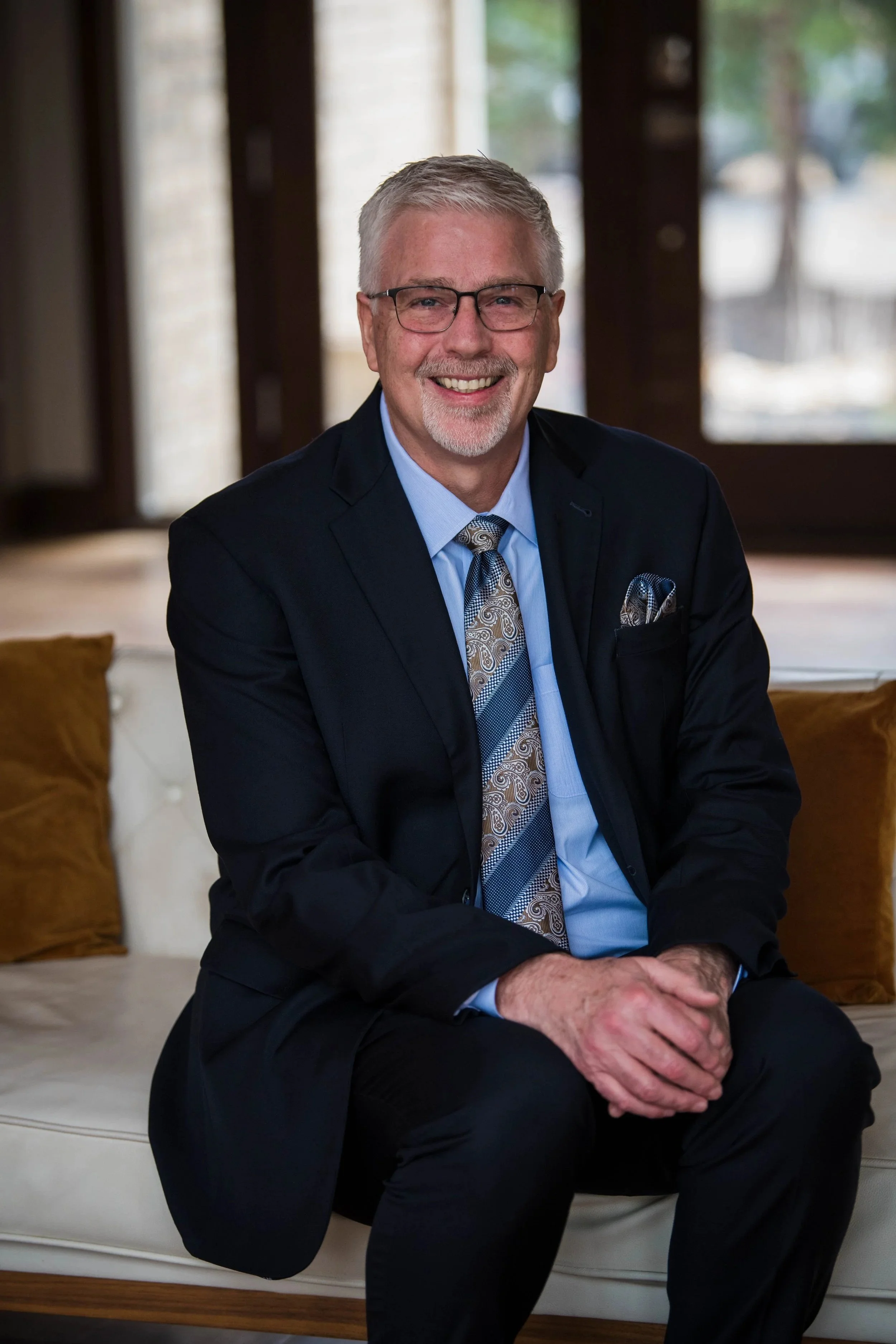 A middle-aged man with gray hair, glasses, and a beard, dressed in a dark suit, blue shirt, and paisley tie, sitting on a cream-colored sofa with brown and beige pillows, smiling at the camera inside a room with large windows showing a blurry outdoor scene.