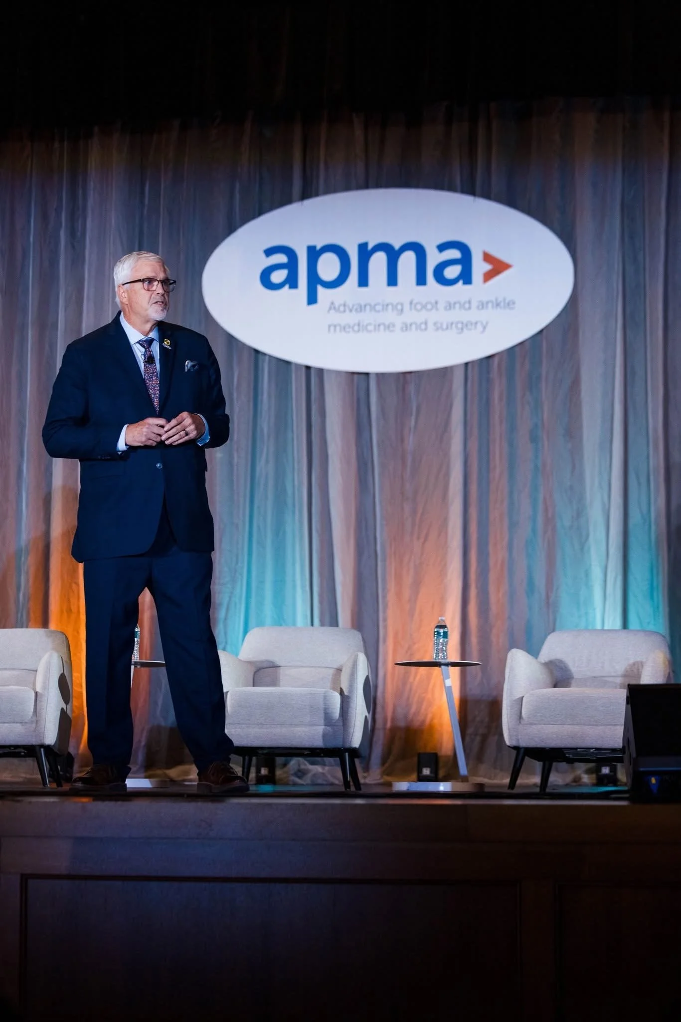 A man with gray hair and glasses standing on a stage with a backdrop that displays the APMA logo and the phrase "Advancing foot and ankle medicine and surgery." There are four empty chairs, small tables with water bottles, and a person is off-screen 