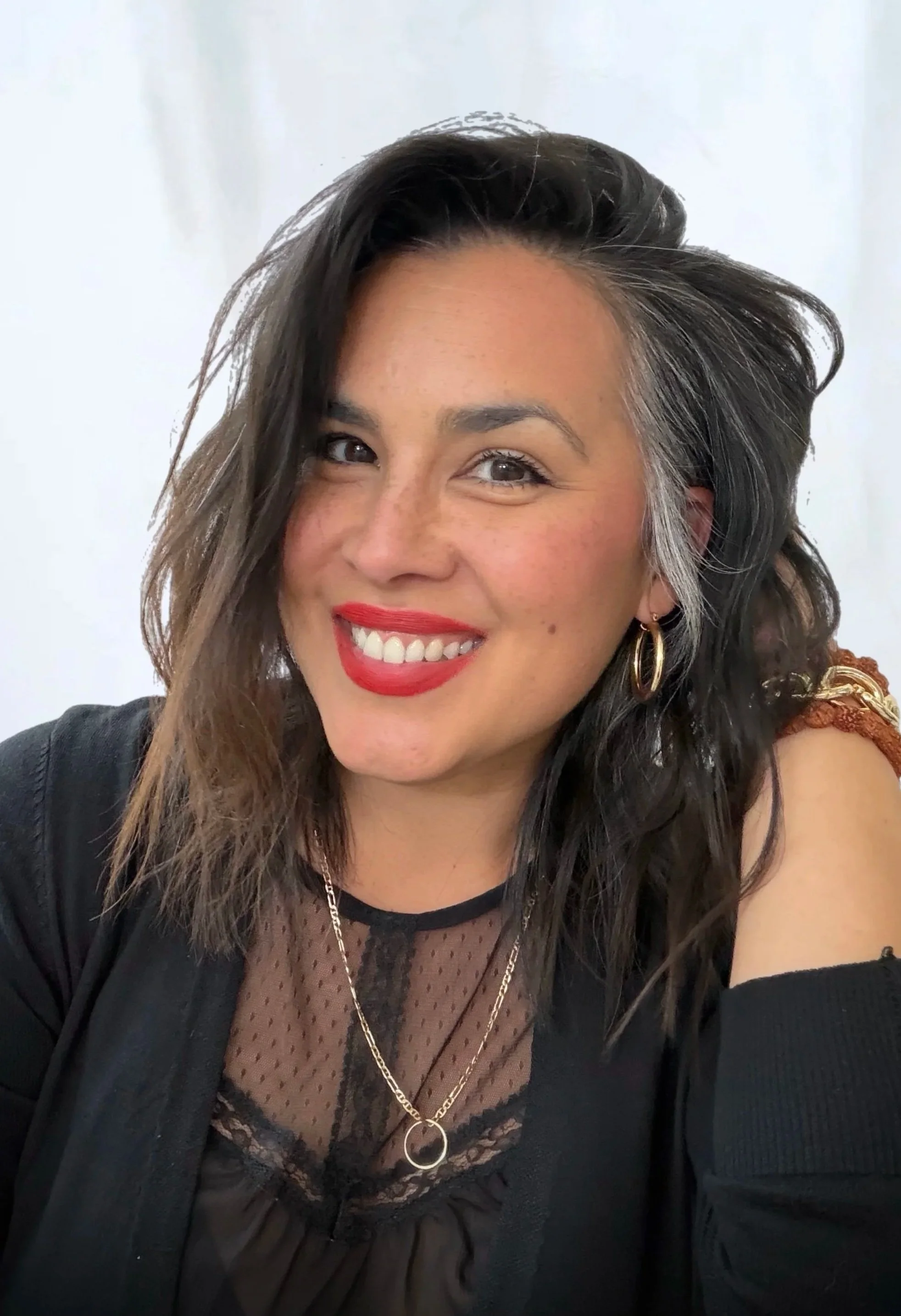 A woman with dark wavy hair wearing a black and metallic patterned top looking at the camera against a light marble background.