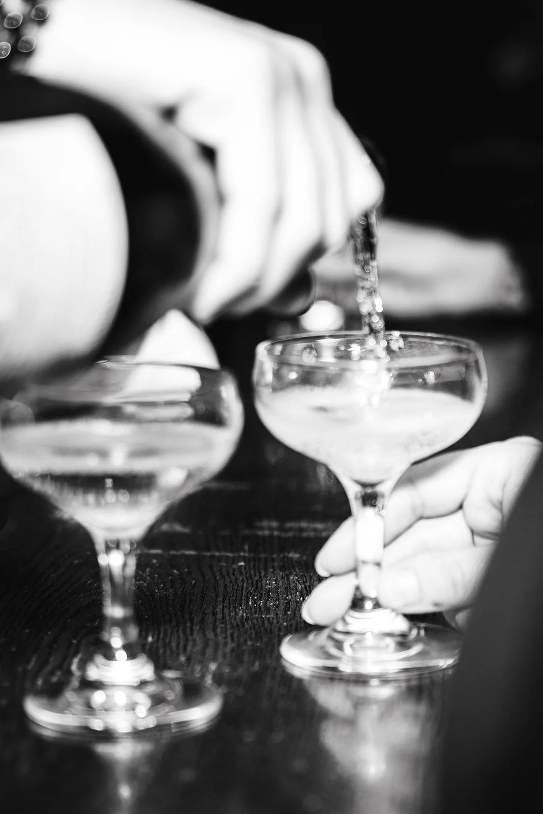 A person pouring a clear drink into a coupe glass at a table, with two similar glasses in the foreground, black and white photo.