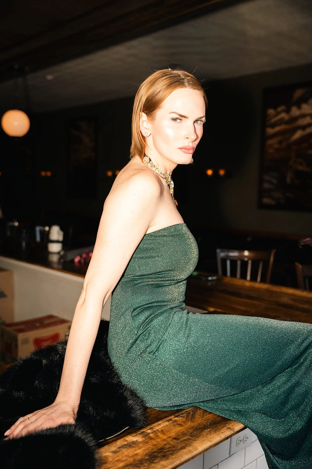 A woman with red hair sitting on a wooden surface in a dimly lit room, wearing a green strapless dress and a statement necklace.