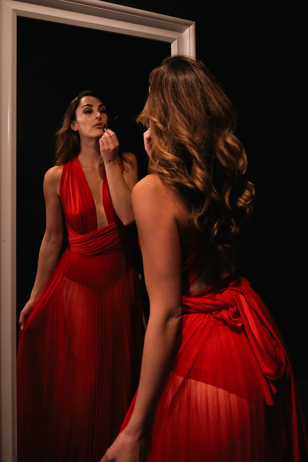A woman in a red gown applying lipstick while looking in a mirror.