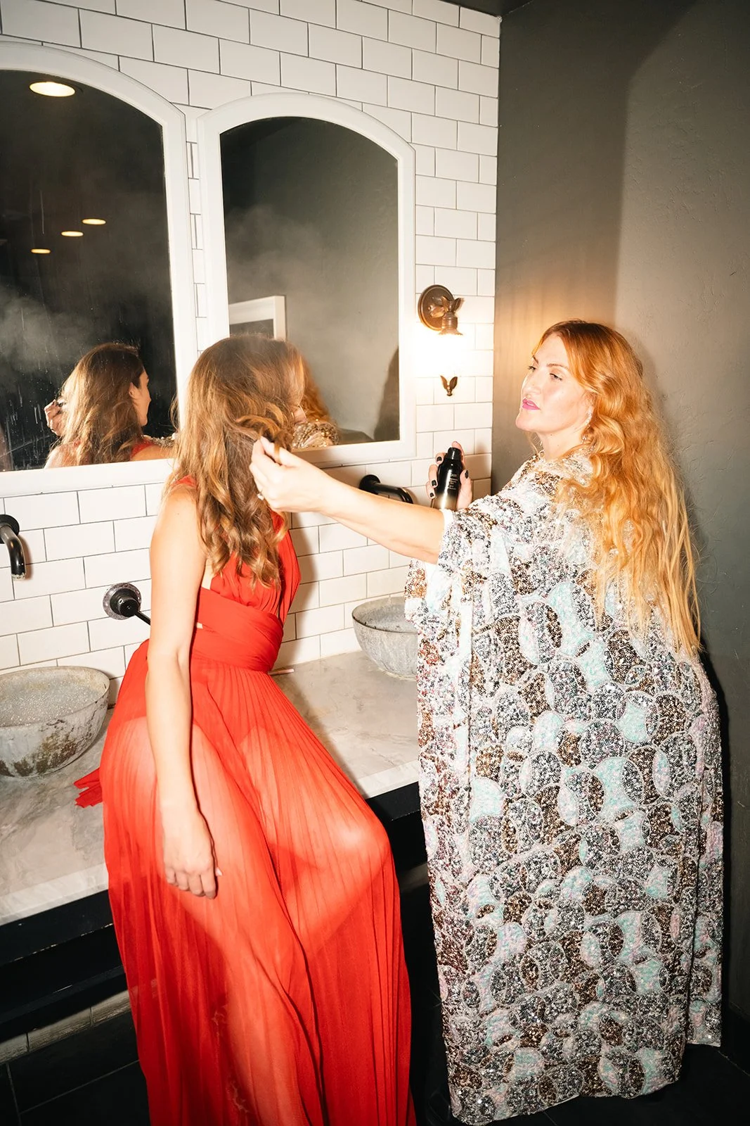 A woman with long, wavy red hair is spray styling another woman with long, curly brown hair, who is wearing a red dress, in a bathroom with white subway tiles and a large mirror.