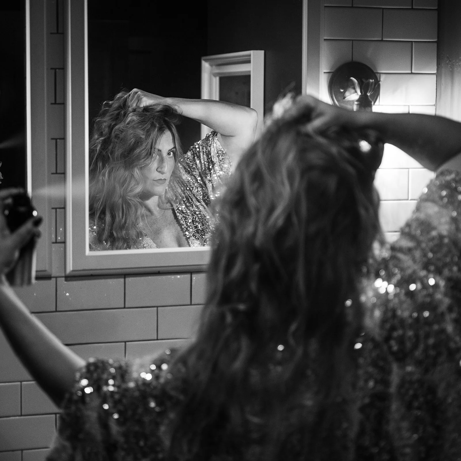 A woman with wavy hair taking a selfie in front of a mirror, with her reflection visible. She is wearing a sequined top and appears to be getting ready, possibly in a bathroom or dressing room.