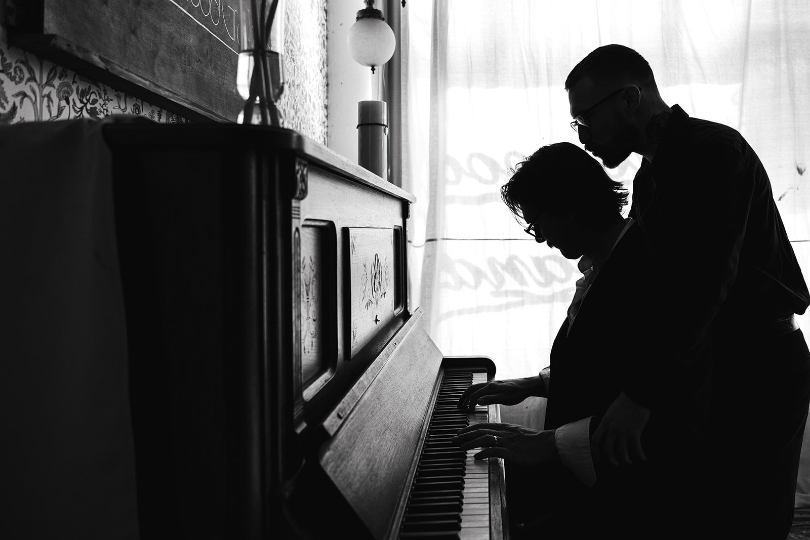 Silhouetted of two people, a man and a woman, playing a piano together in a room with curtains.