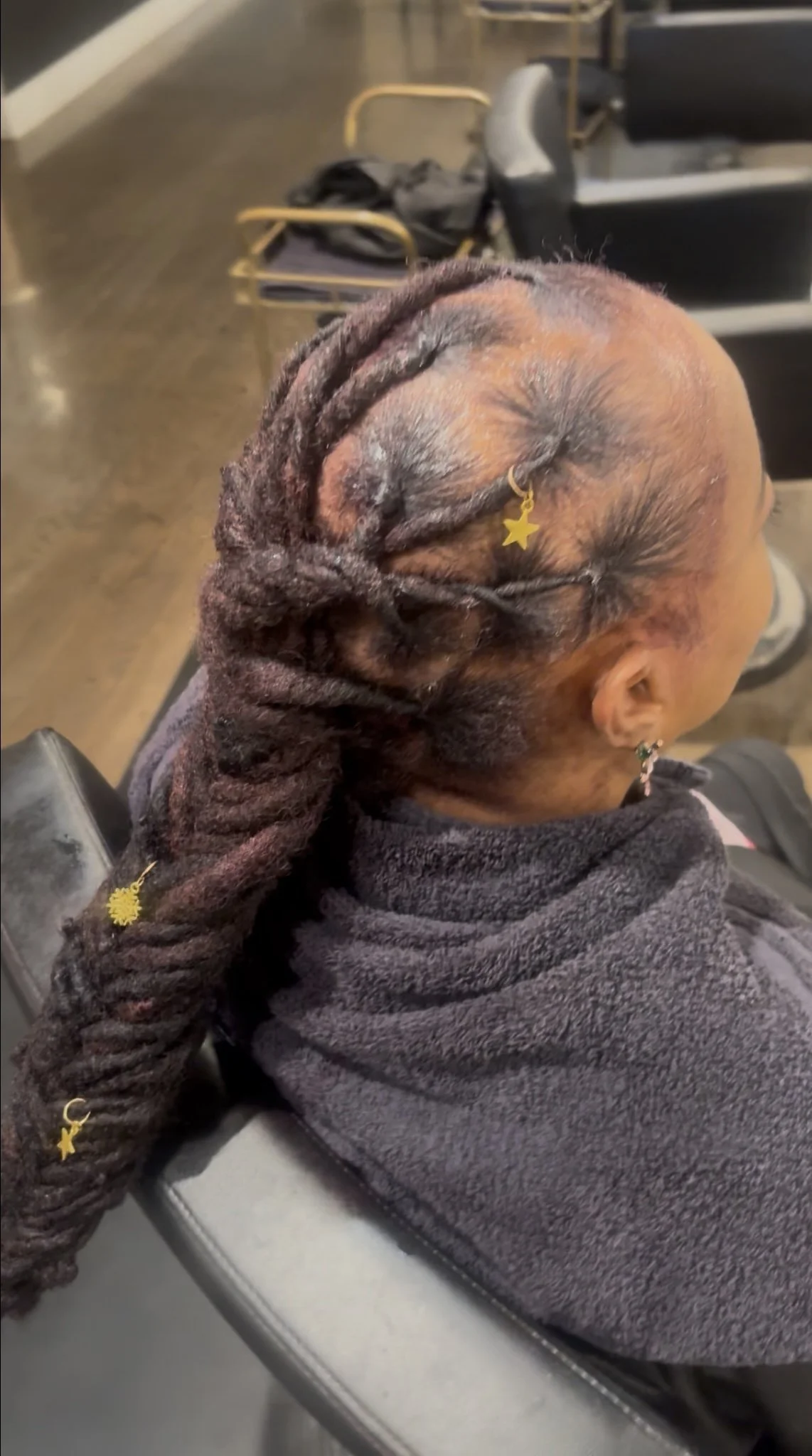 A woman with long dreadlocks decorated with gold star charms, sitting in a salon chair, with visible scalp and hair treatment.