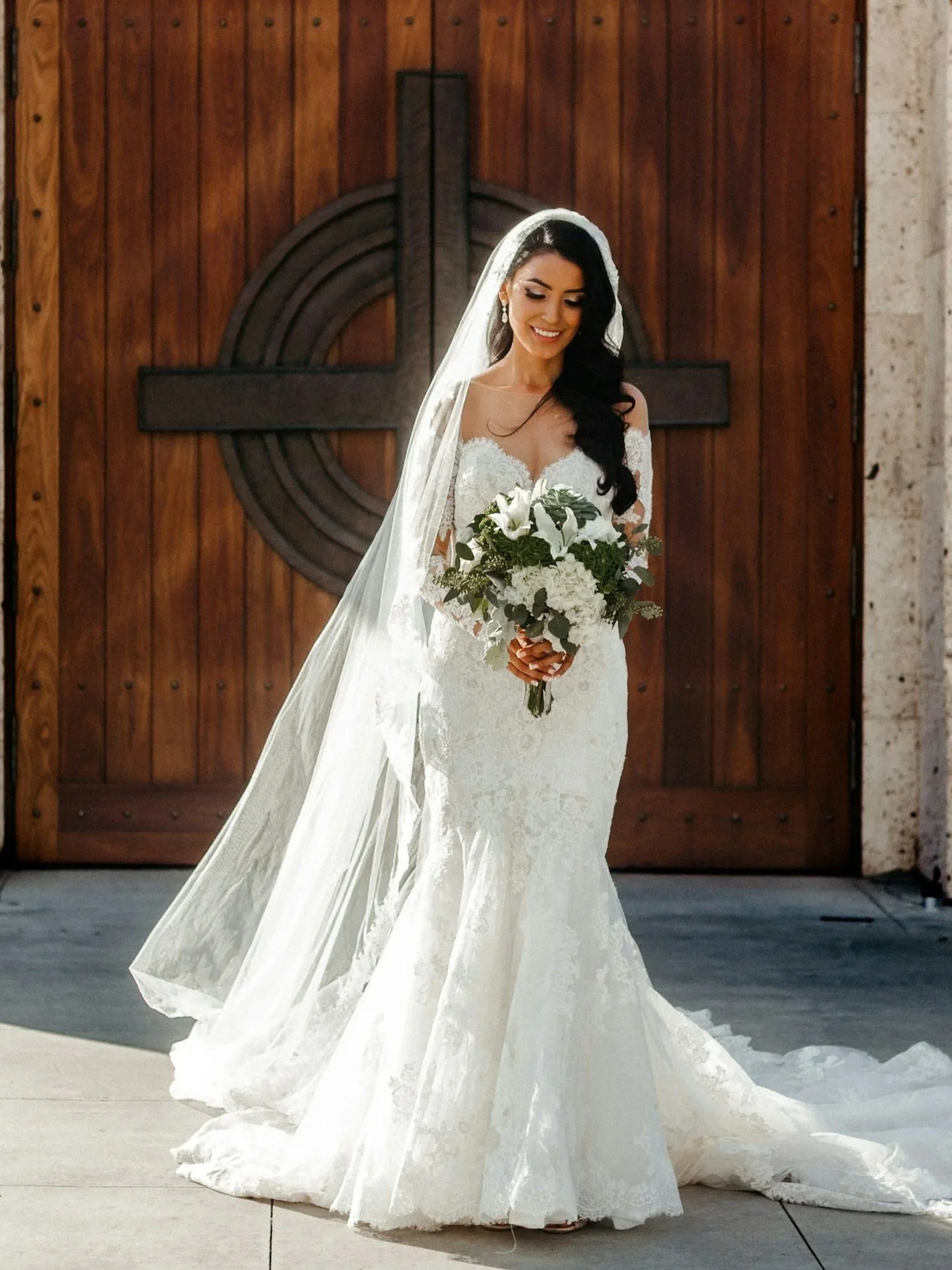 Smiling bride in a white wedding gown holding a bouquet of white flowers, standing in front of a wooden door with a large cross design.