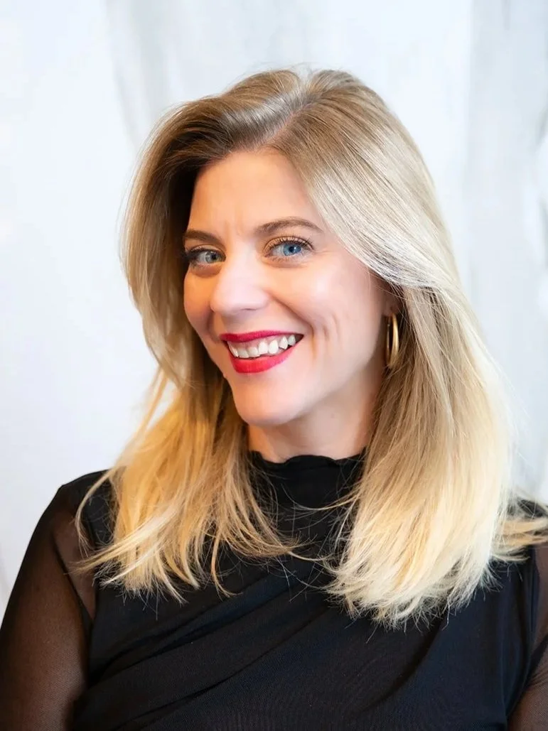 A woman with blonde hair, blue eyes, and red lipstick smiling, wearing gold hoop earrings and a black top, standing against a light background.
