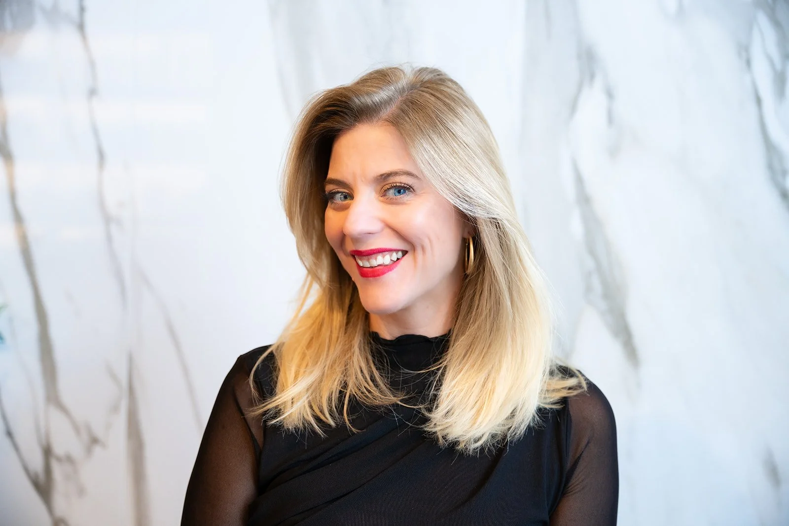 A smiling woman with blonde hair, blue eyes, wearing a black top and gold hoop earrings, standing in front of a light-colored, textured wall.