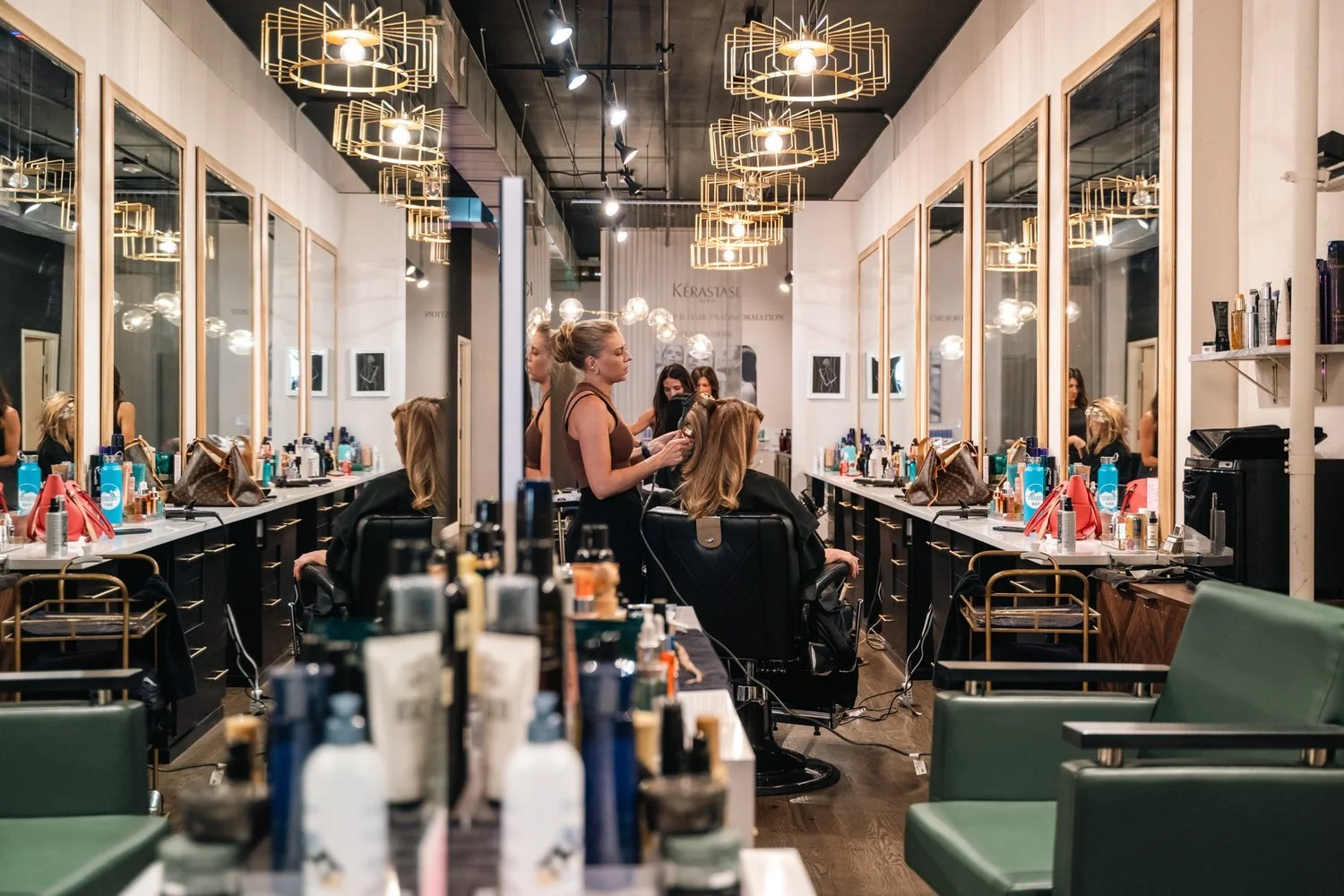Interior of a modern hair salon with clients getting hair styled, mirrors, hair products on counters, salon chairs, and pendant lighting.