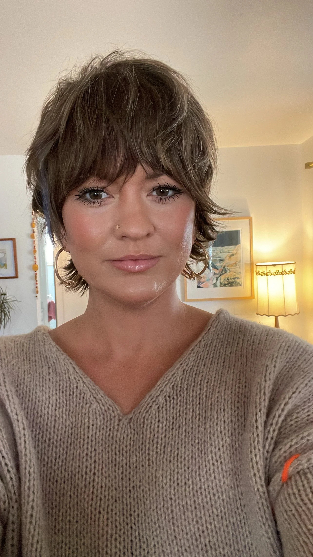 A woman with short, wavy brown hair and makeup, wearing a beige sweater, taking a selfie indoors with a lamp and framed artwork in the background.