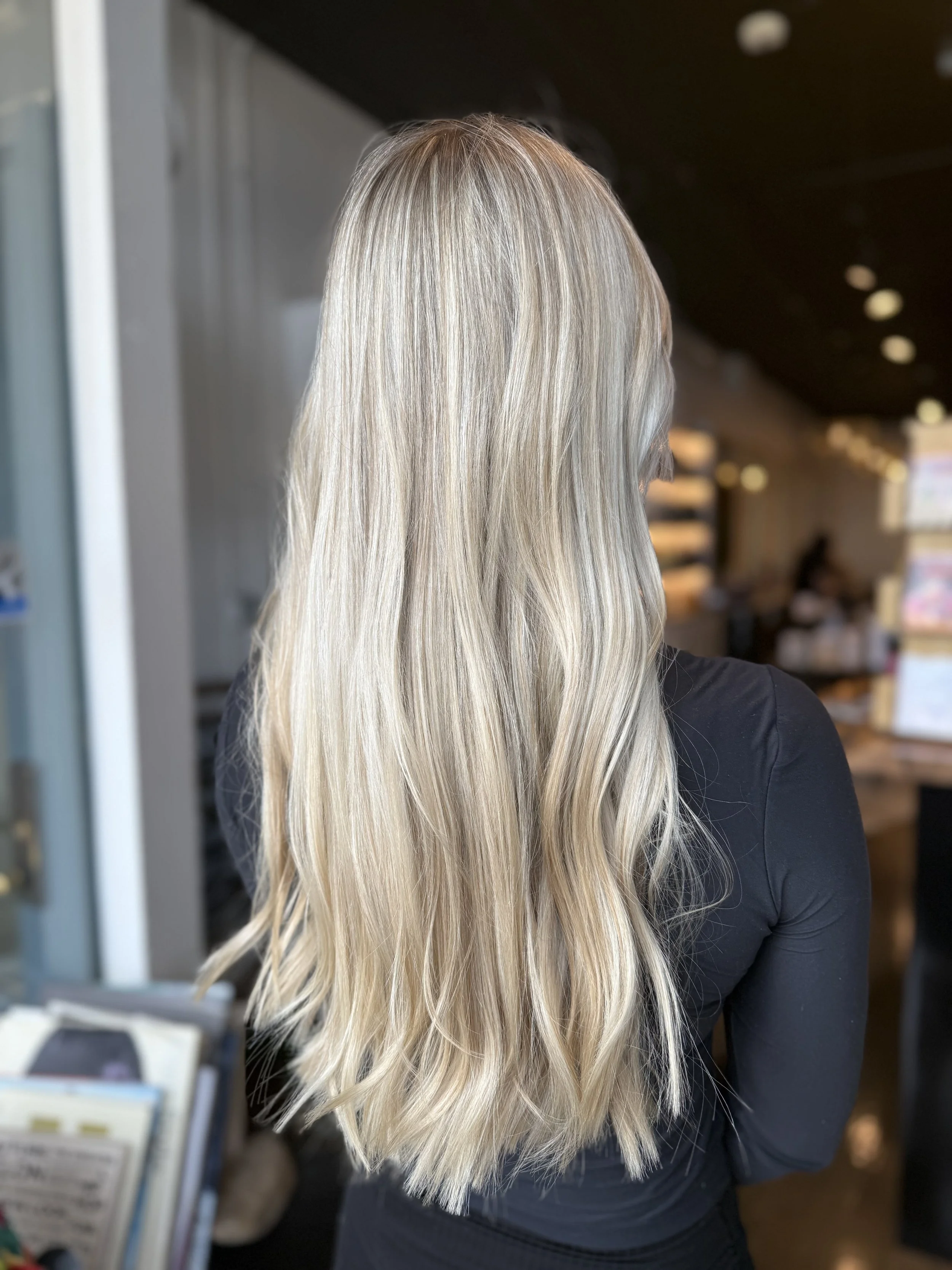 Blonde woman with wavy hair wearing a black top, photographed from behind in an indoor setting.