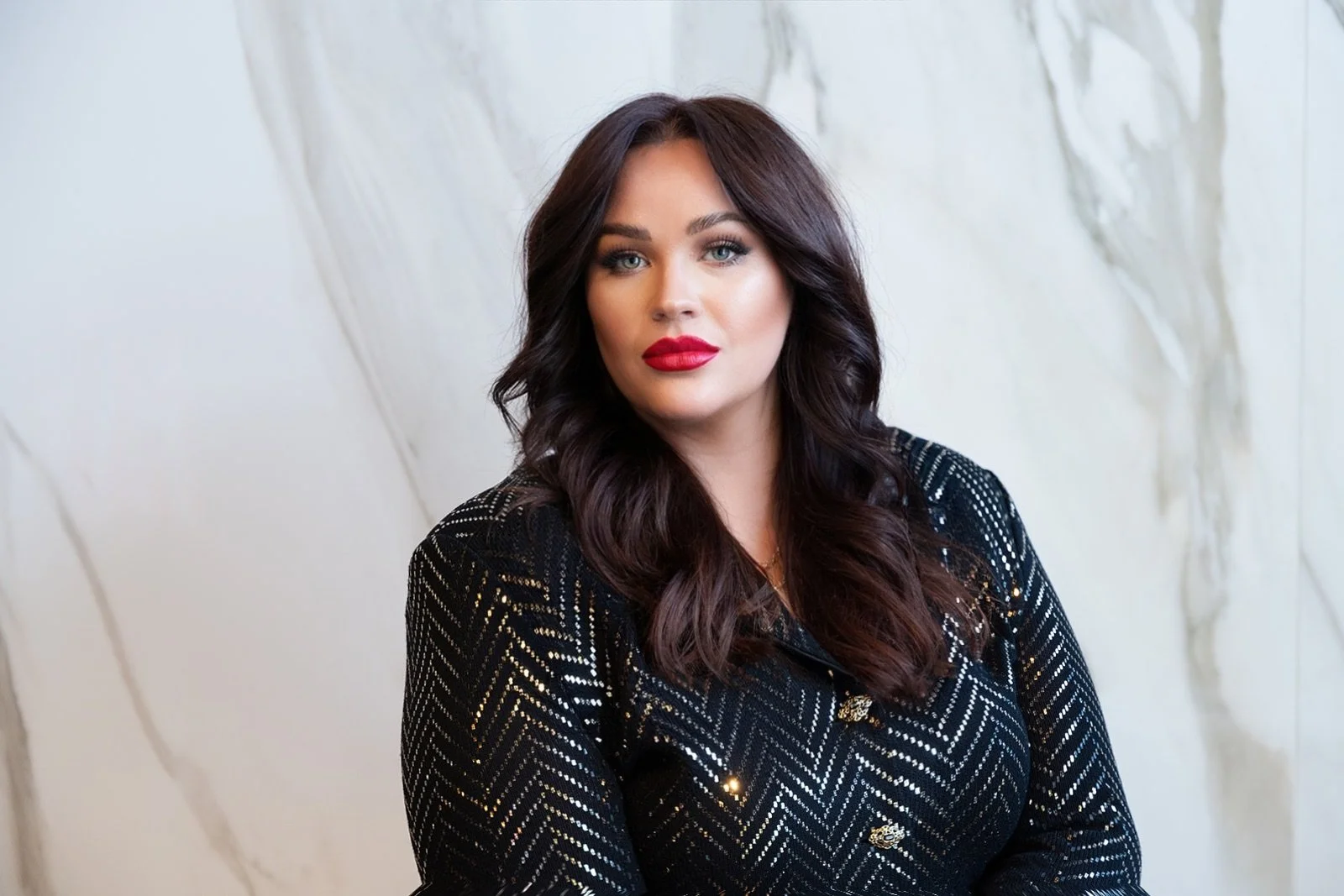 A woman with dark wavy hair wearing a black and metallic patterned top looking at the camera against a light marble background.