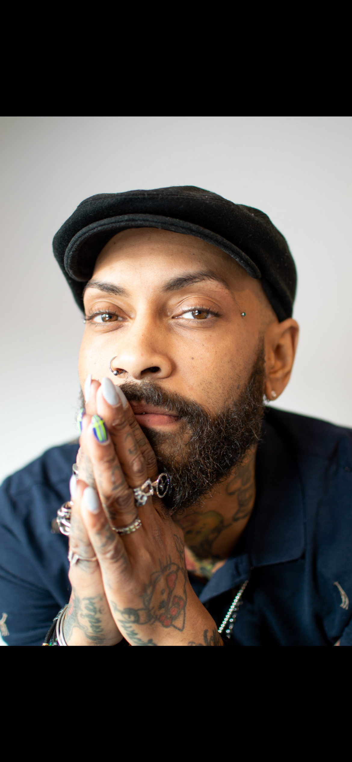 Close-up portrait of a man with a beard, tattoos, piercings, and multiple rings and rings on fingers, wearing a black beret, with hand partially covering his mouth against a plain, light-colored background.