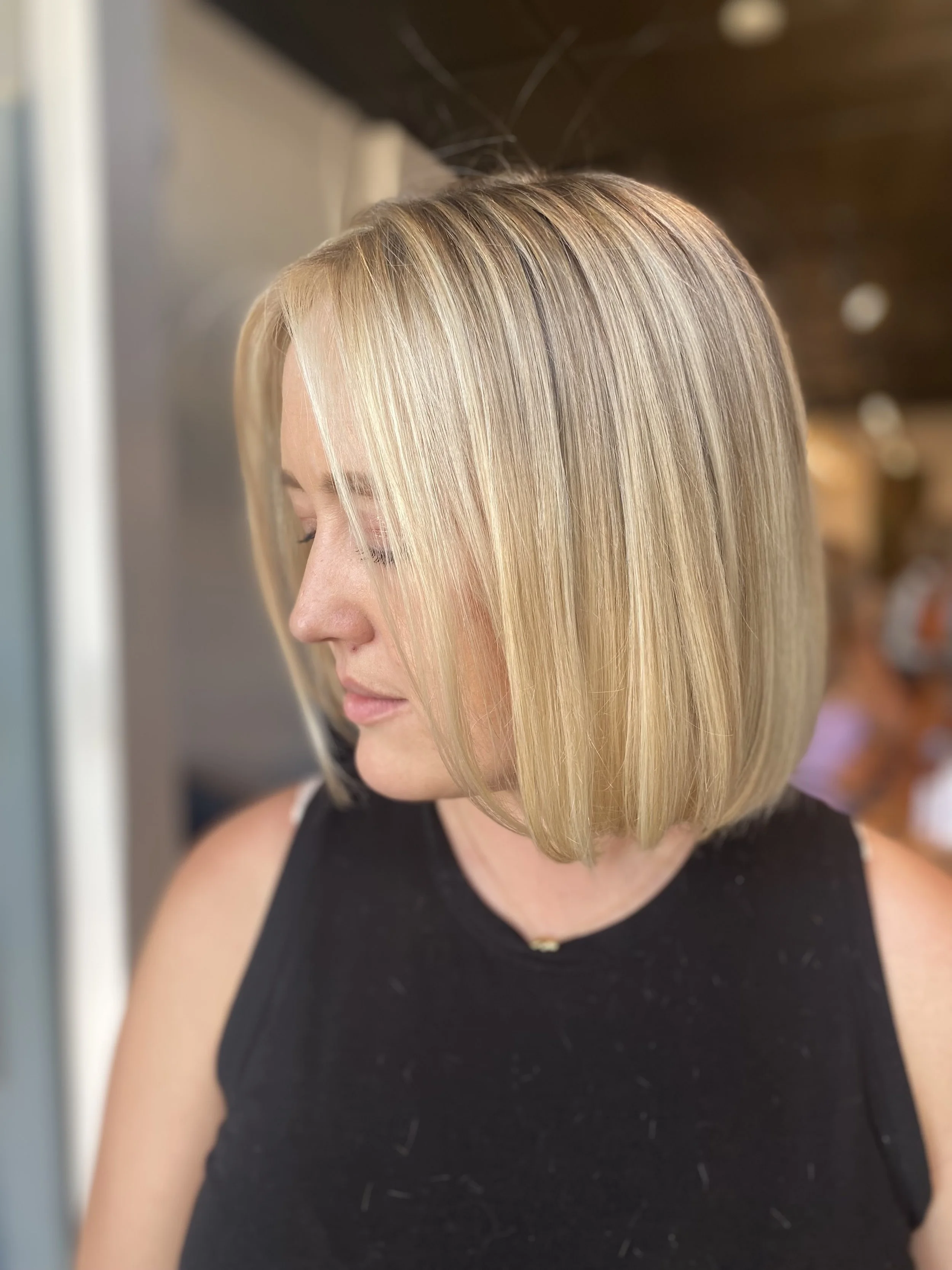 Side view of a woman with a blonde bob haircut, looking downward, in a black sleeveless top, indoors.