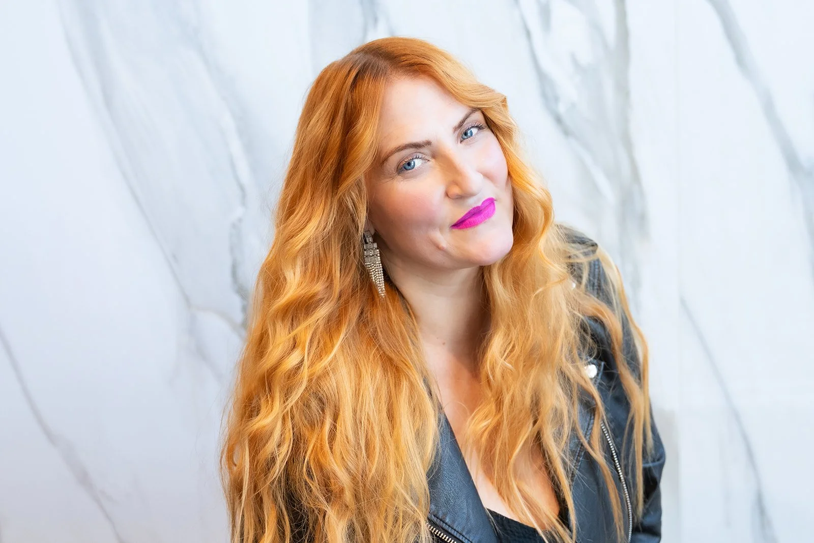 A woman with long red hair, wearing a black leather jacket and large earrings, standing against a white marble background.