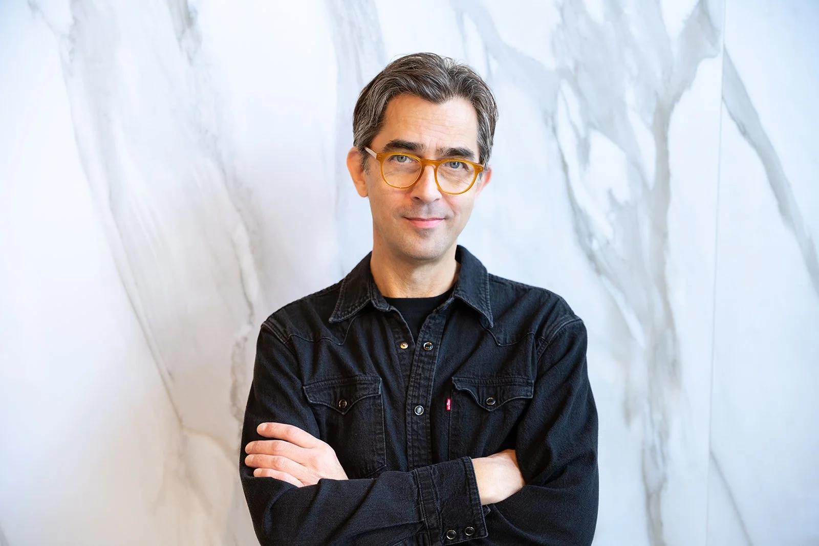A man with gray hair and glasses, wearing a black denim jacket, crossing his arms and standing in front of a white marble wall.