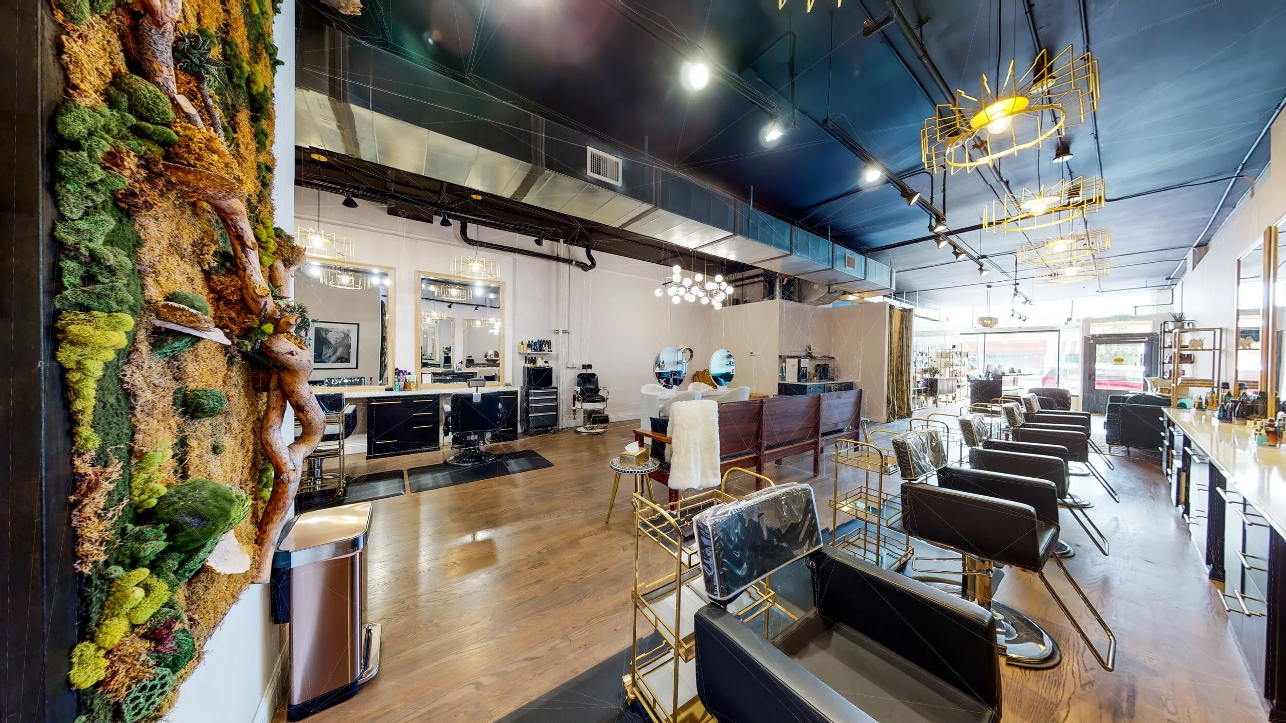 Interior view of a modern hair salon with black salon chairs, gold accents, wooden flooring, and large mirrors, illuminated by ceiling lights and decorative chandeliers.