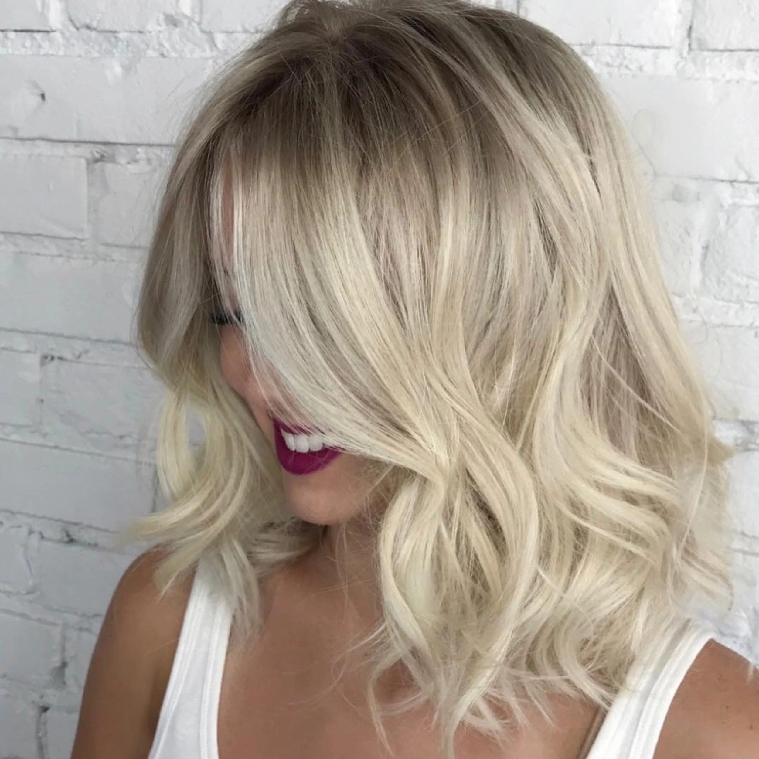 A woman with blonde, wavy bob haircut, wearing dark lipstick, smiling, against a white brick wall.