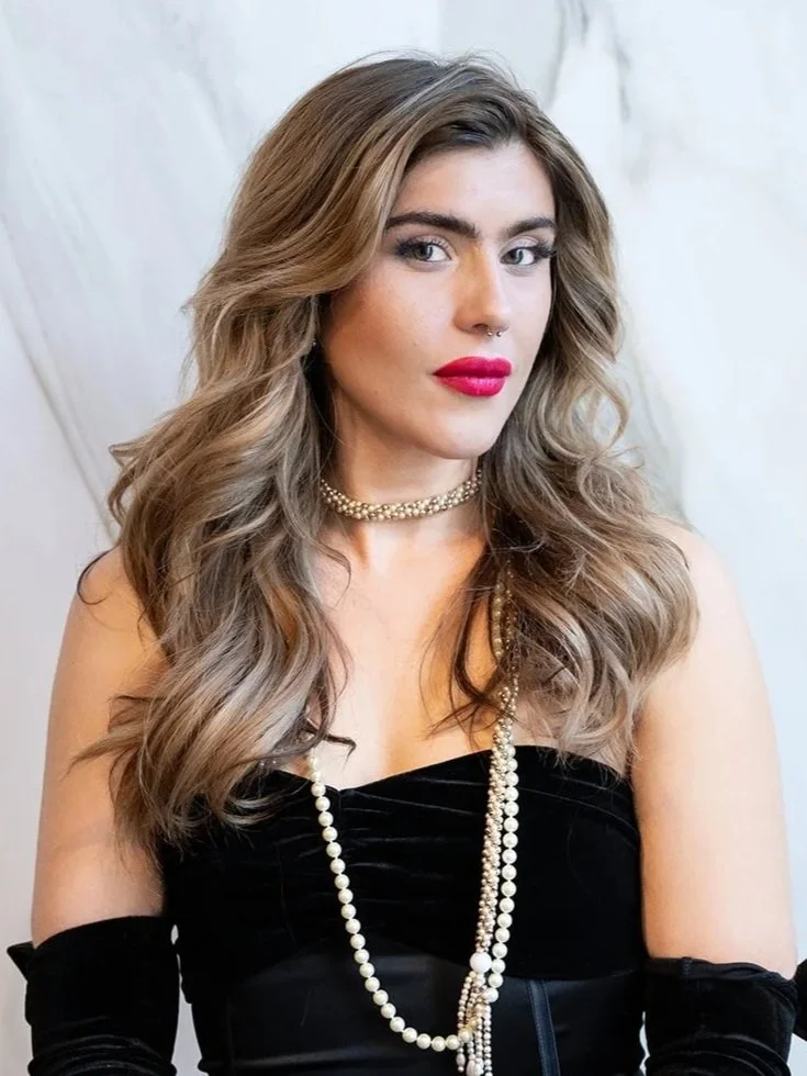 A woman with long wavy blonde hair, wearing bright pink lipstick, a nose ring, a pearl choker, and layered pearl necklaces, dressed in a black outfit with off-shoulder sleeves, standing against a light-colored background.