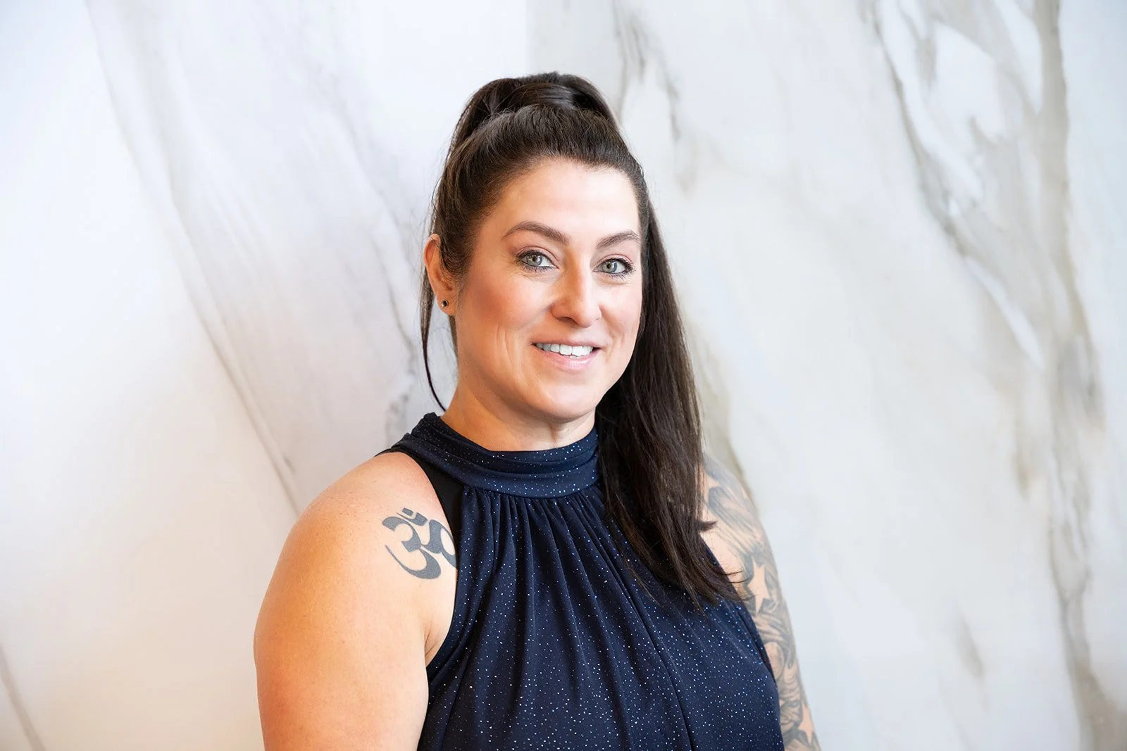 A woman with long dark hair tied up in a high ponytail, wearing a navy sleeveless top with a sparkly pattern, standing against a textured white wall. She has tattoos on her right shoulder and arm, including an Om symbol. She is smiling and looking at the camera.