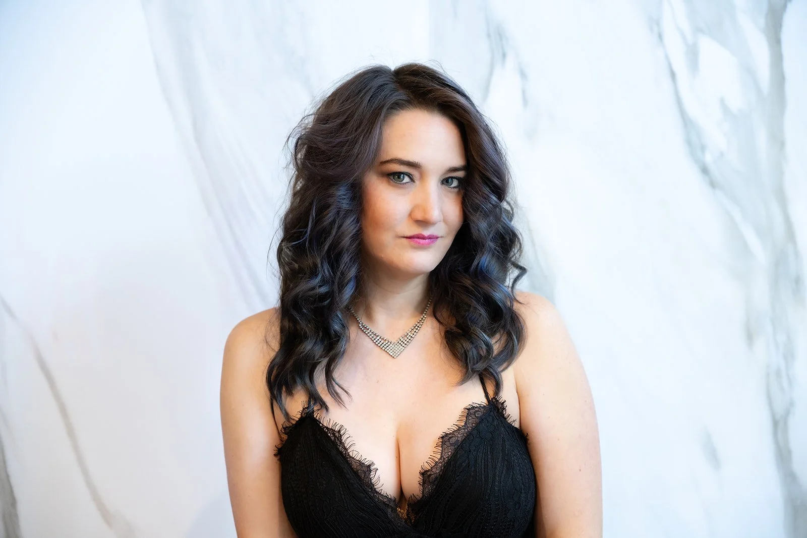 A woman with dark wavy hair wearing a black lace dress and a silver necklace, standing in front of a white textured wall.