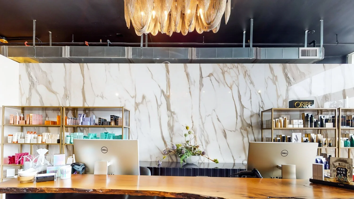 Interior of a reception or checkout area with a marble wall, shelves of beauty products, and two Dell monitors on a wooden counter, with a flower arrangement in the center.