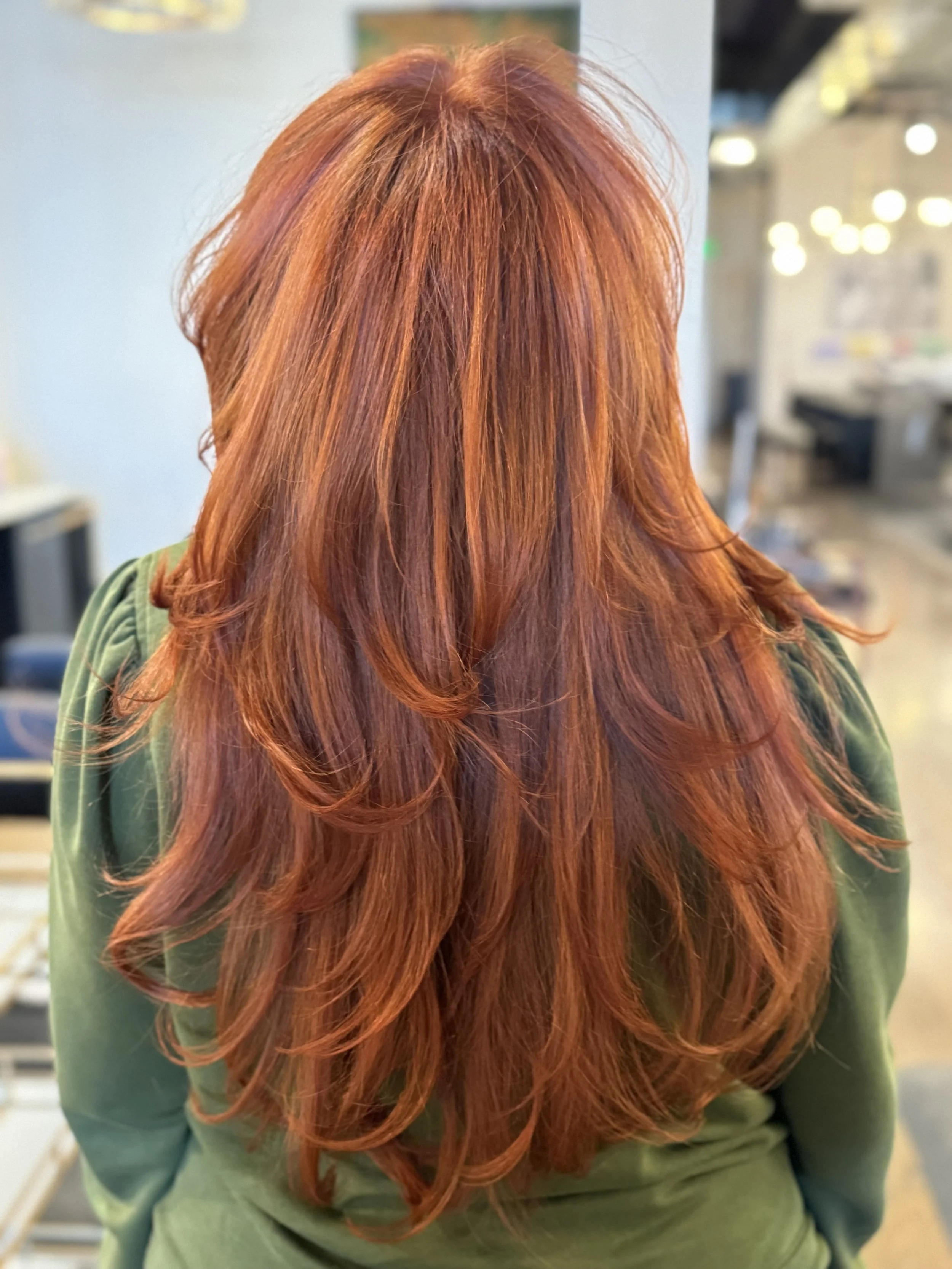 Back of a woman with long, wavy, reddish-orange hair wearing a green top in a salon.