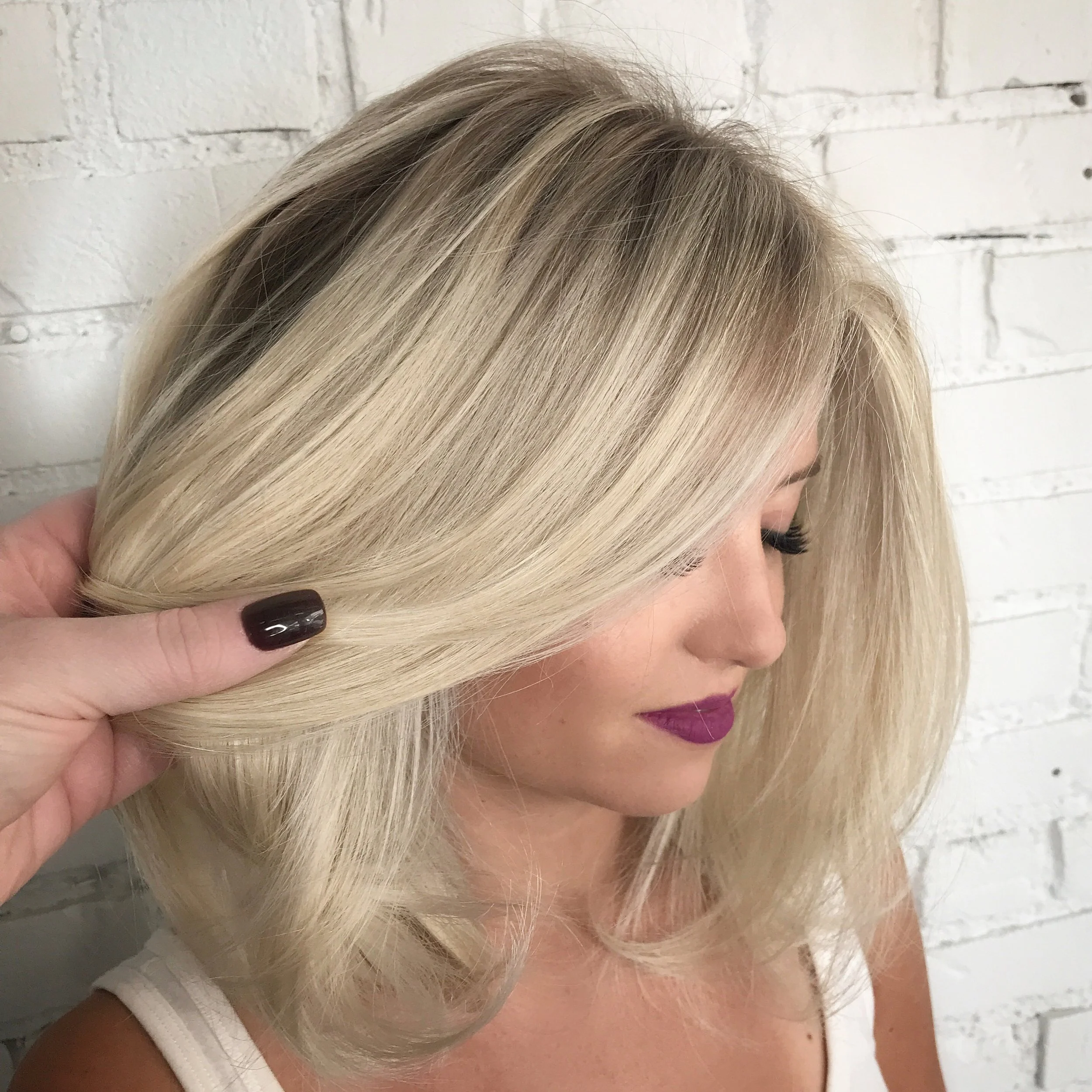 Close-up of a woman with platinum blonde hair and dark lipstick, with a hand holding a section of her hair. The background is a white brick wall.