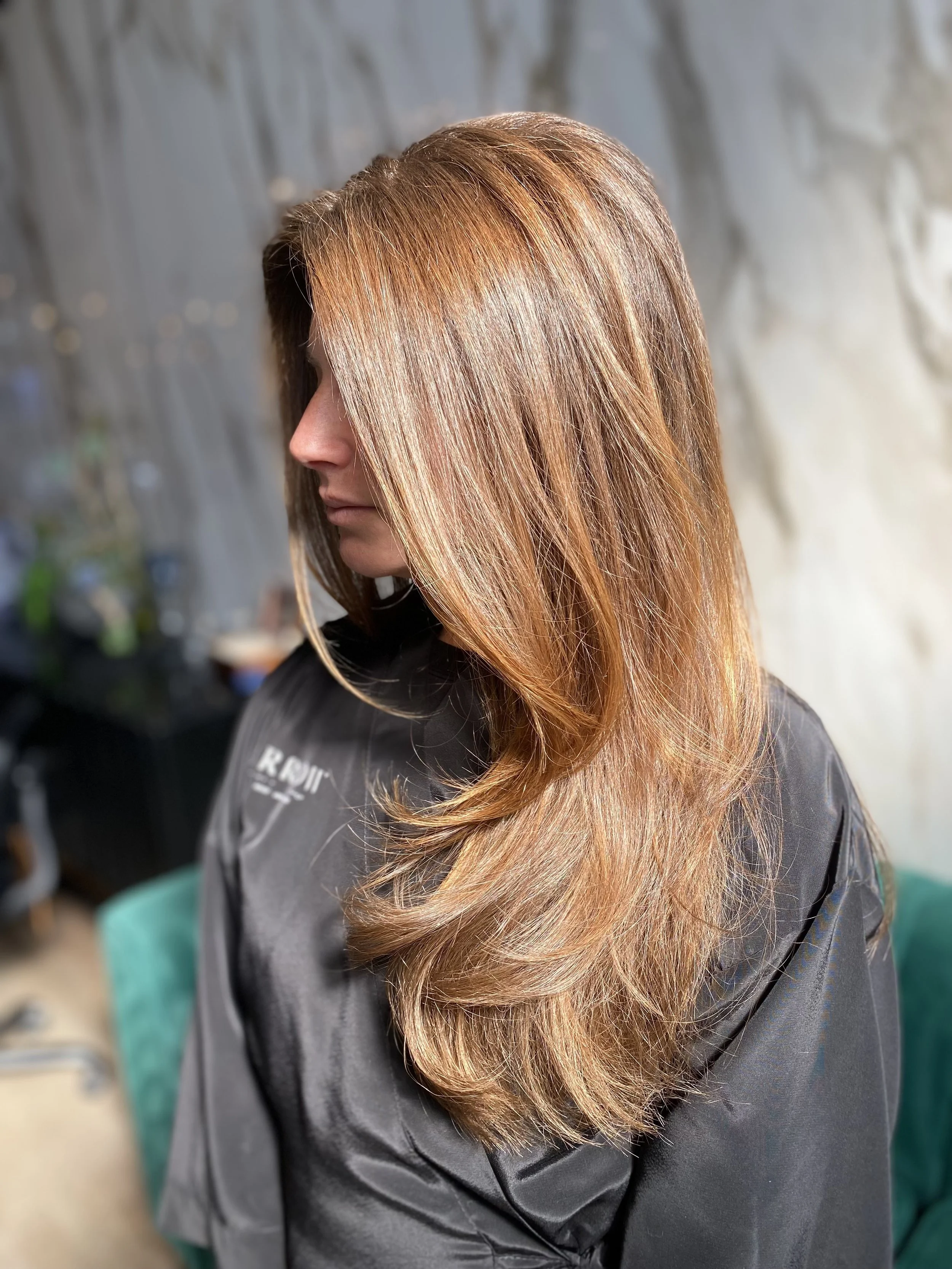 Side profile of a woman with long, layered, reddish-brown hair, wearing a black salon cape.