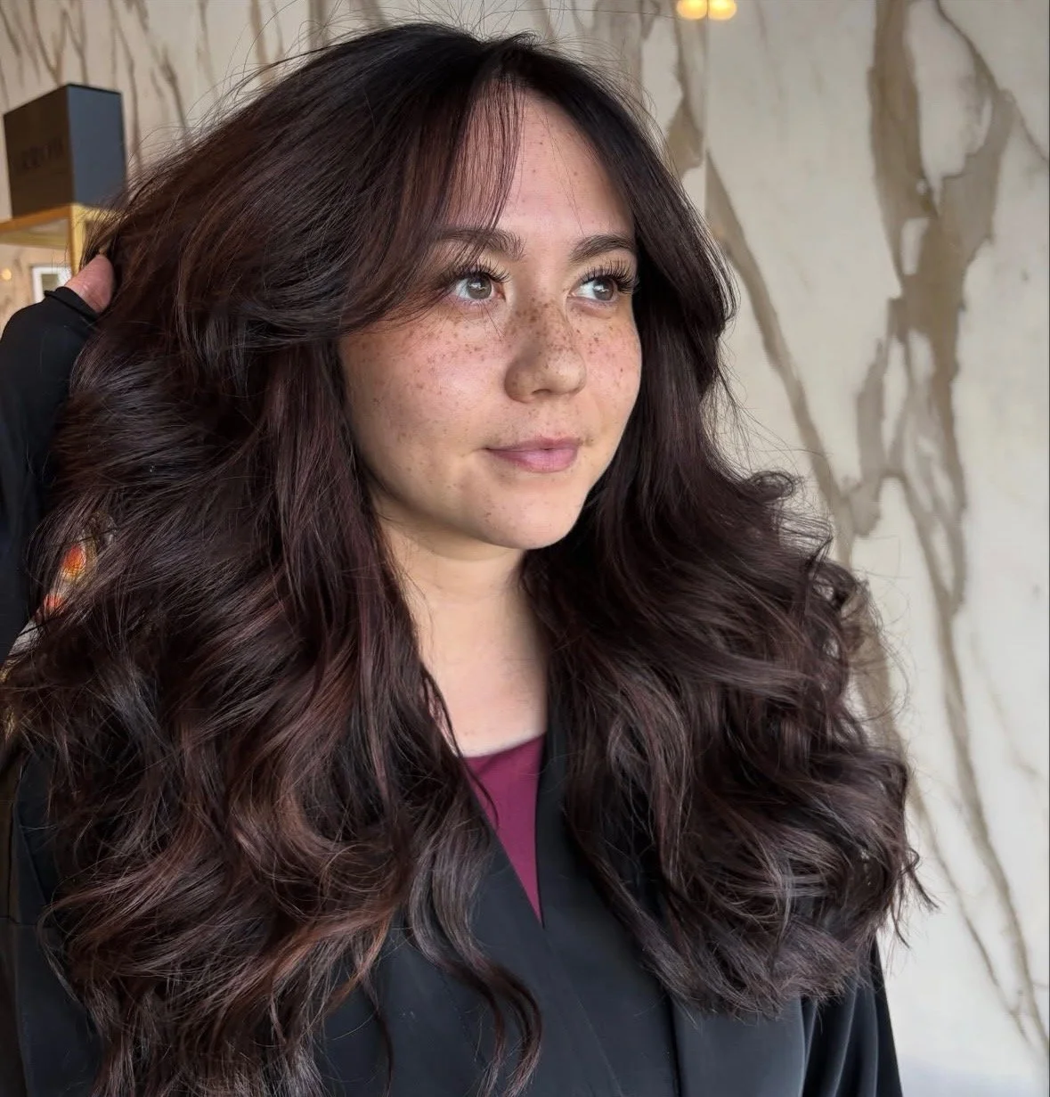 A woman with long, wavy chestnut hair, freckles, and light-colored eyes, looking slightly to the right, outdoors with a marble wall in the background.