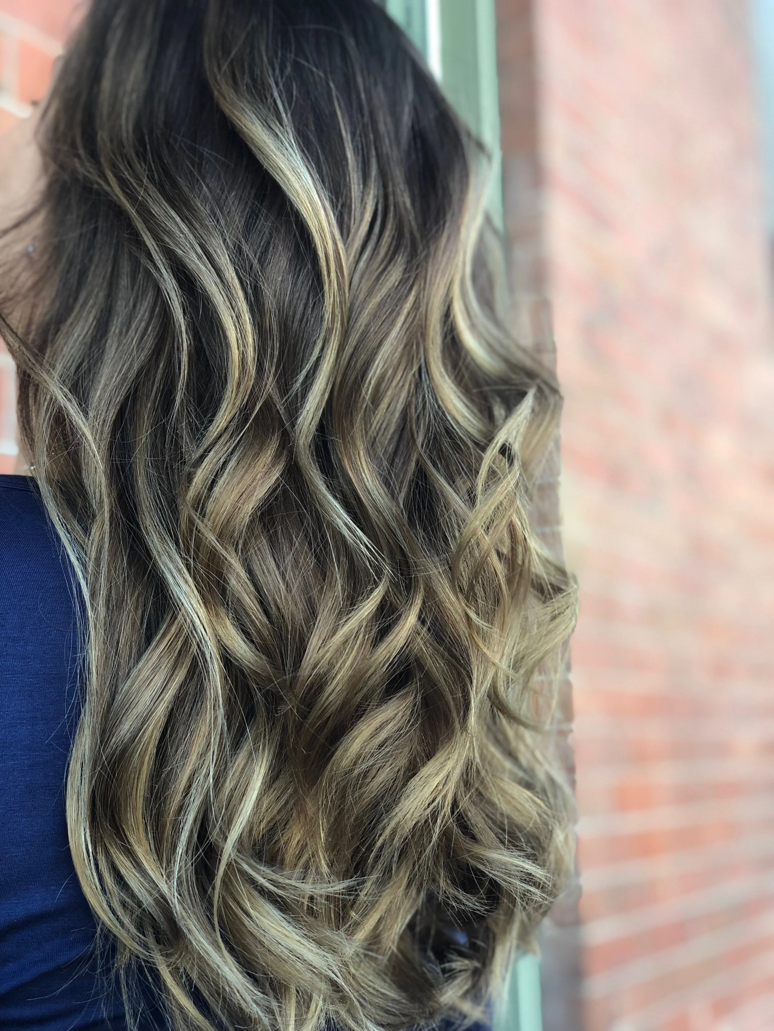 Close-up side view of long, wavy hair with blonde highlights and darker roots, outdoors against a red brick wall.