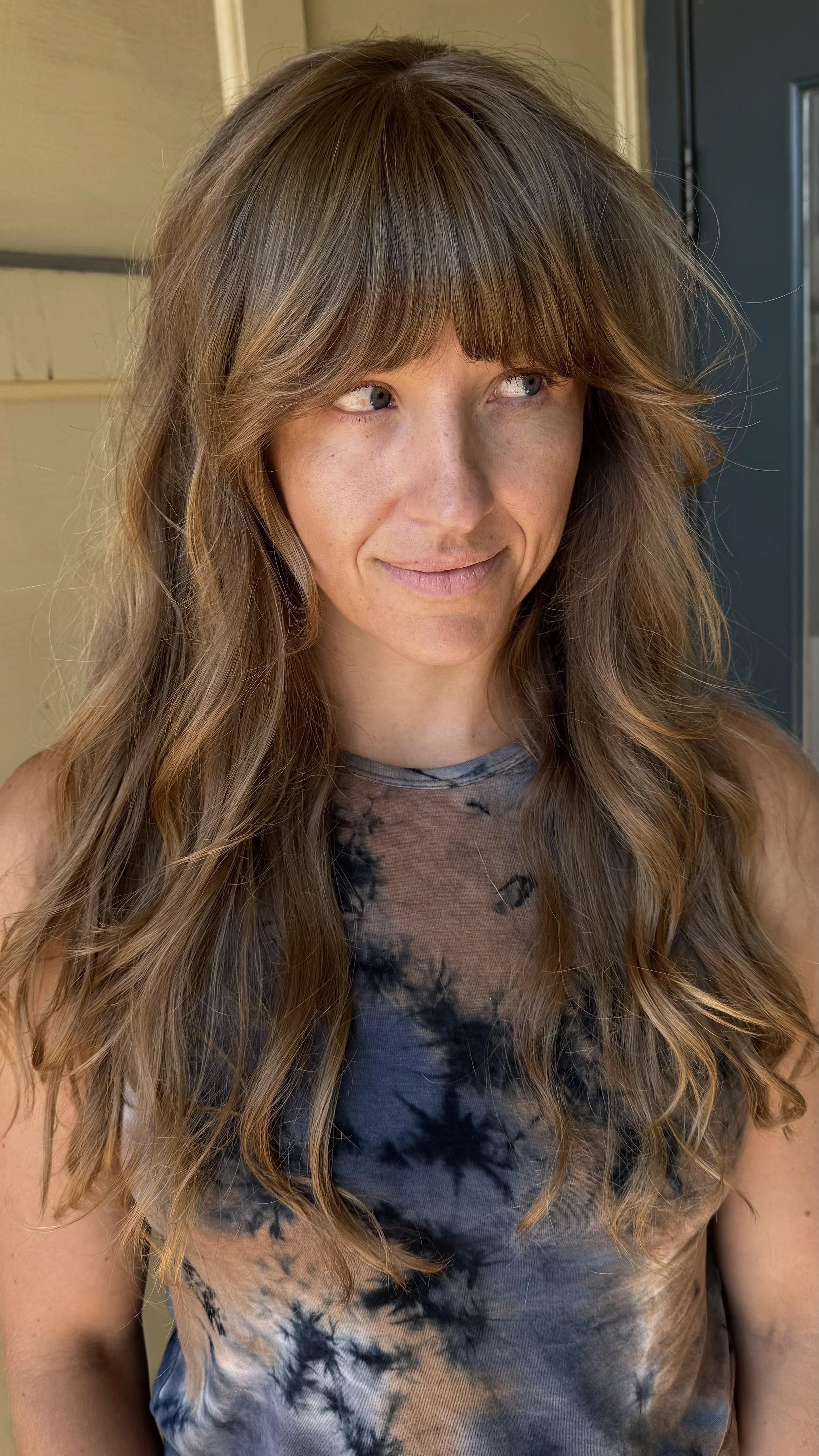A woman with long, wavy light brown hair and bangs, wearing a sleeveless tie-dye top in black and beige, standing outdoors near a house with a beige wall and blue door.