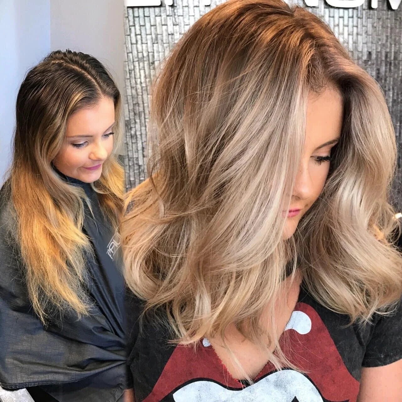 Two women with styled blonde hair looking down, one in the foreground with voluminous hair and a black shirt with a logo, the other in the background with a striped shirt and a black cape, against a textured gray wall.
