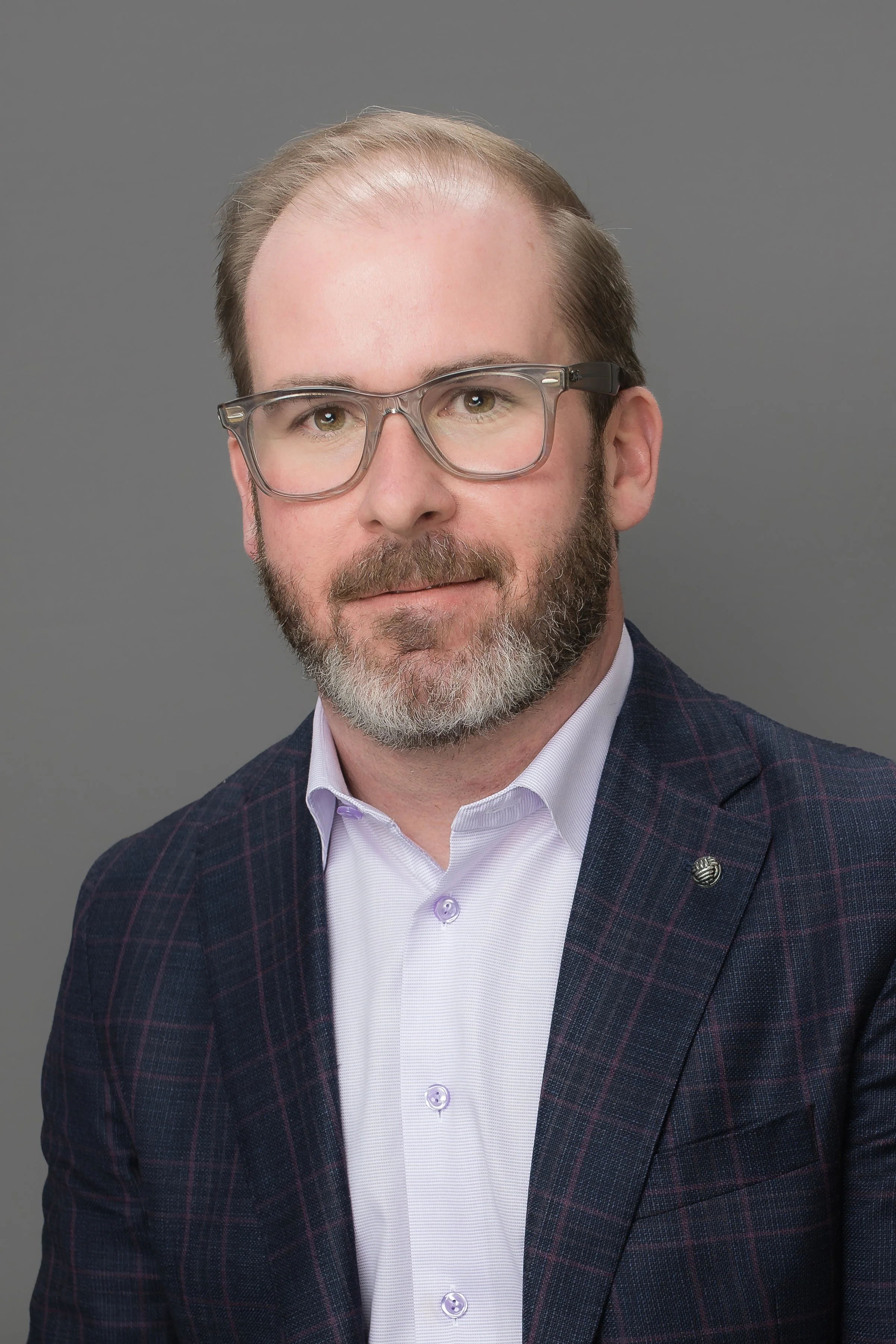 Headshot of a man with glasses, beard, and mustache wearing a checkered blazer and white shirt against a gray background.