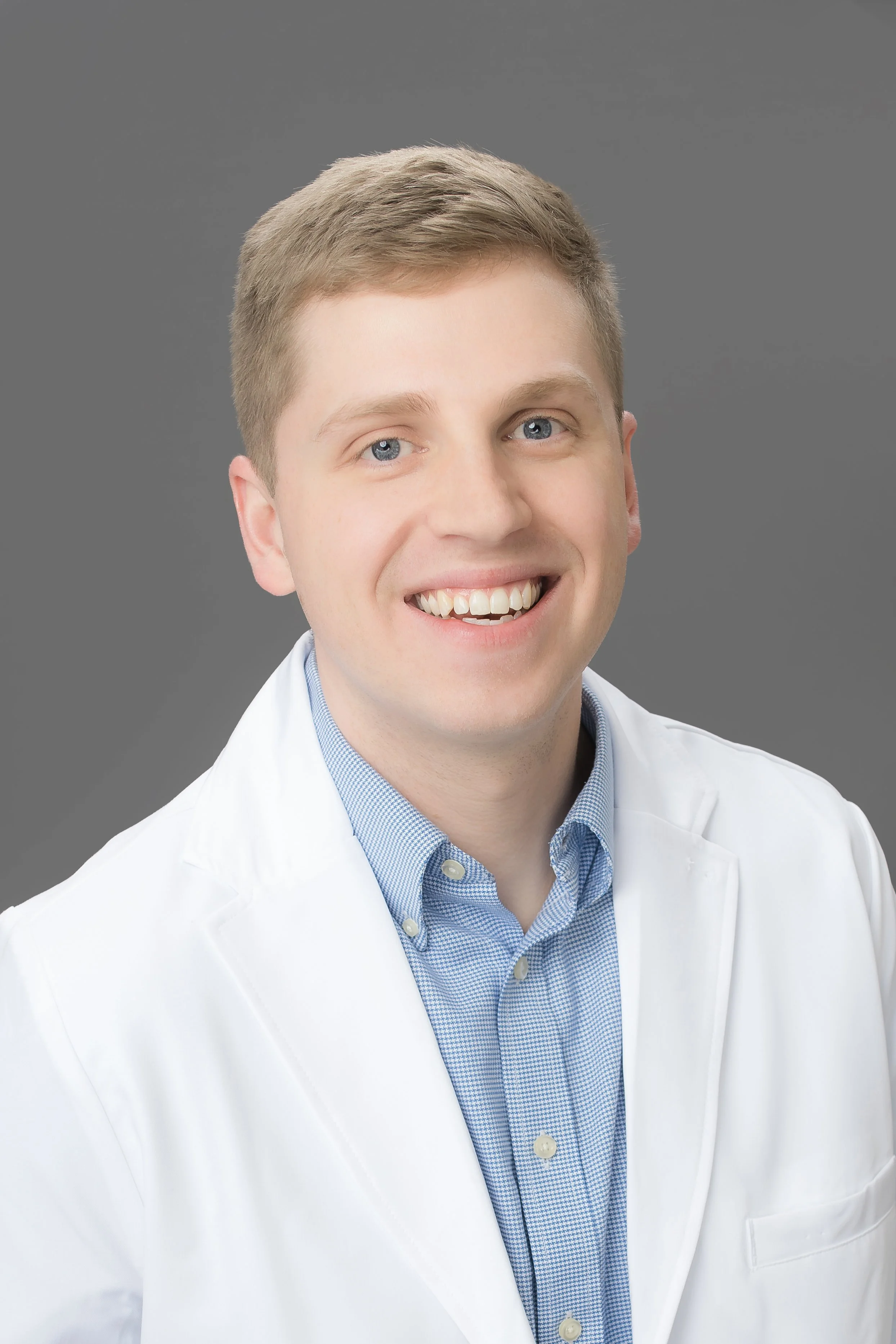 A young male doctor wearing a white coat and blue button-up shirt, smiling with a gray background.